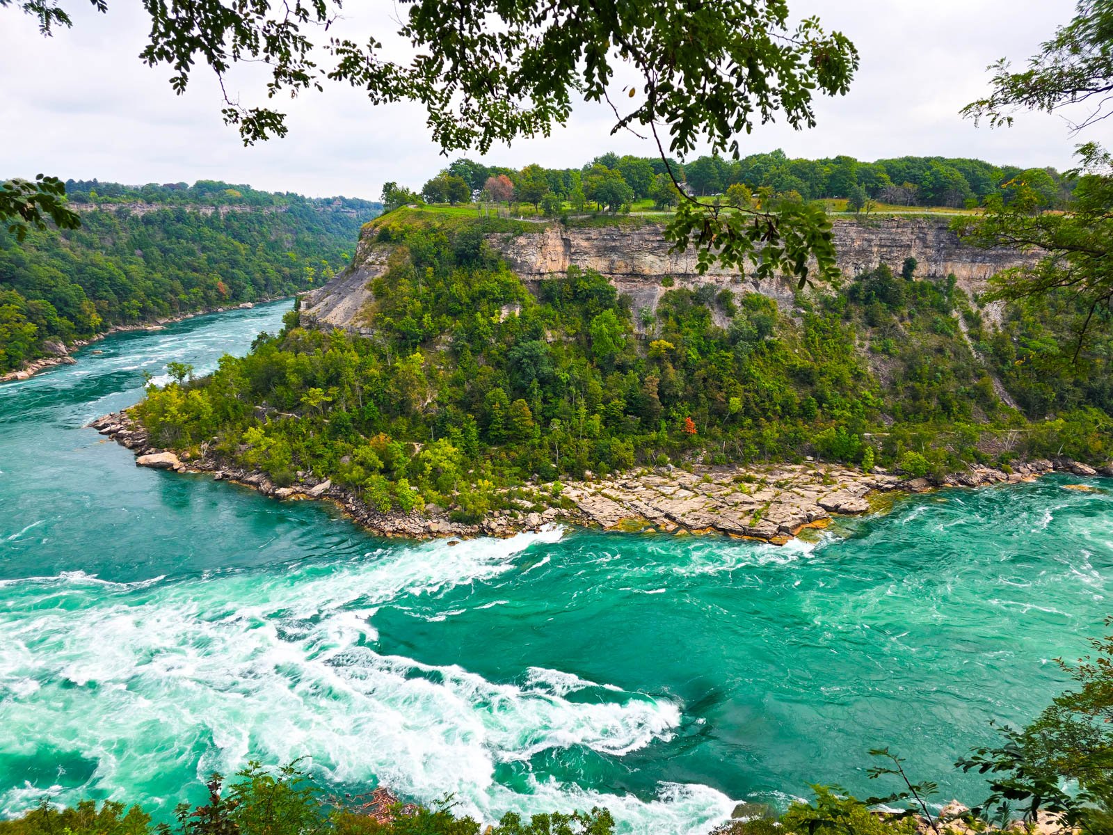 A sharpe fork in a river with rocky cliff faces covered in trees, the river is bright turquoise with white waves on the fast-flowing turn of the river, see from a viewpoint above