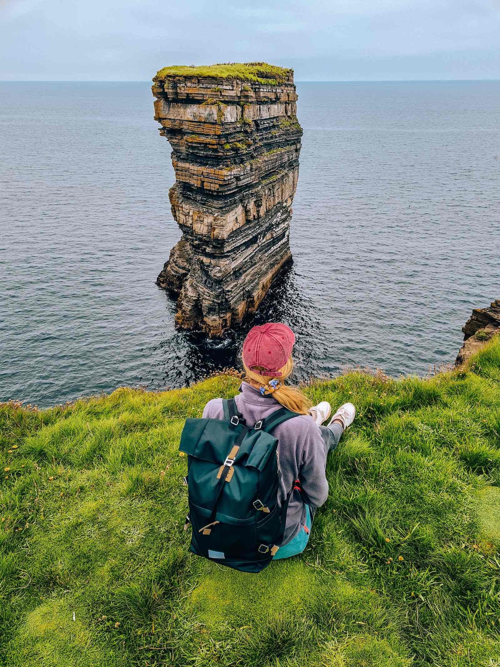 A girl sitting on the edge of a cliff looking out onto a sea stack sticking out of the ocean