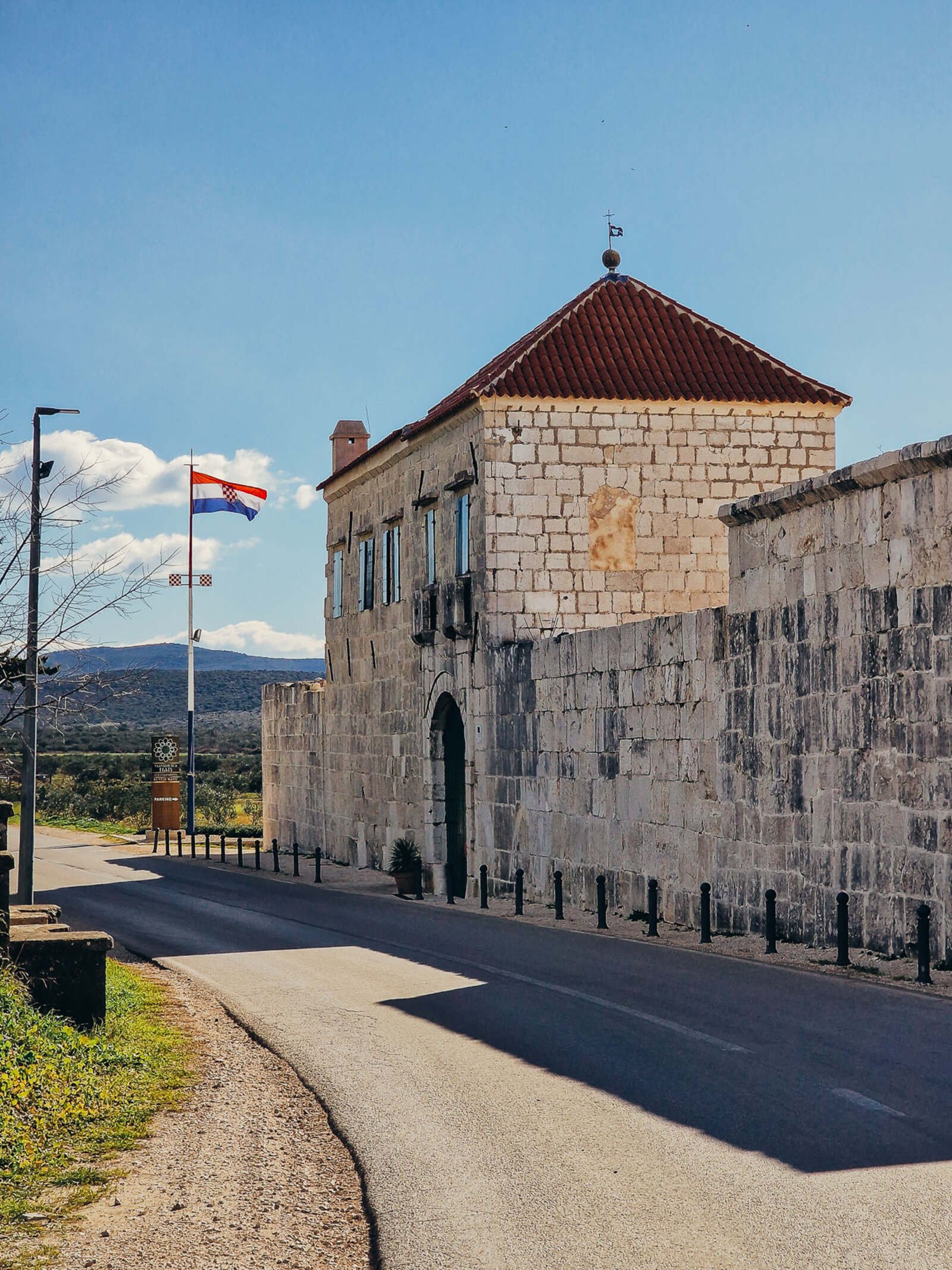 A large square stone building that looks very old with a red tile roof and a Croatian flag flying outside. It's right next to the road and the sky is blue