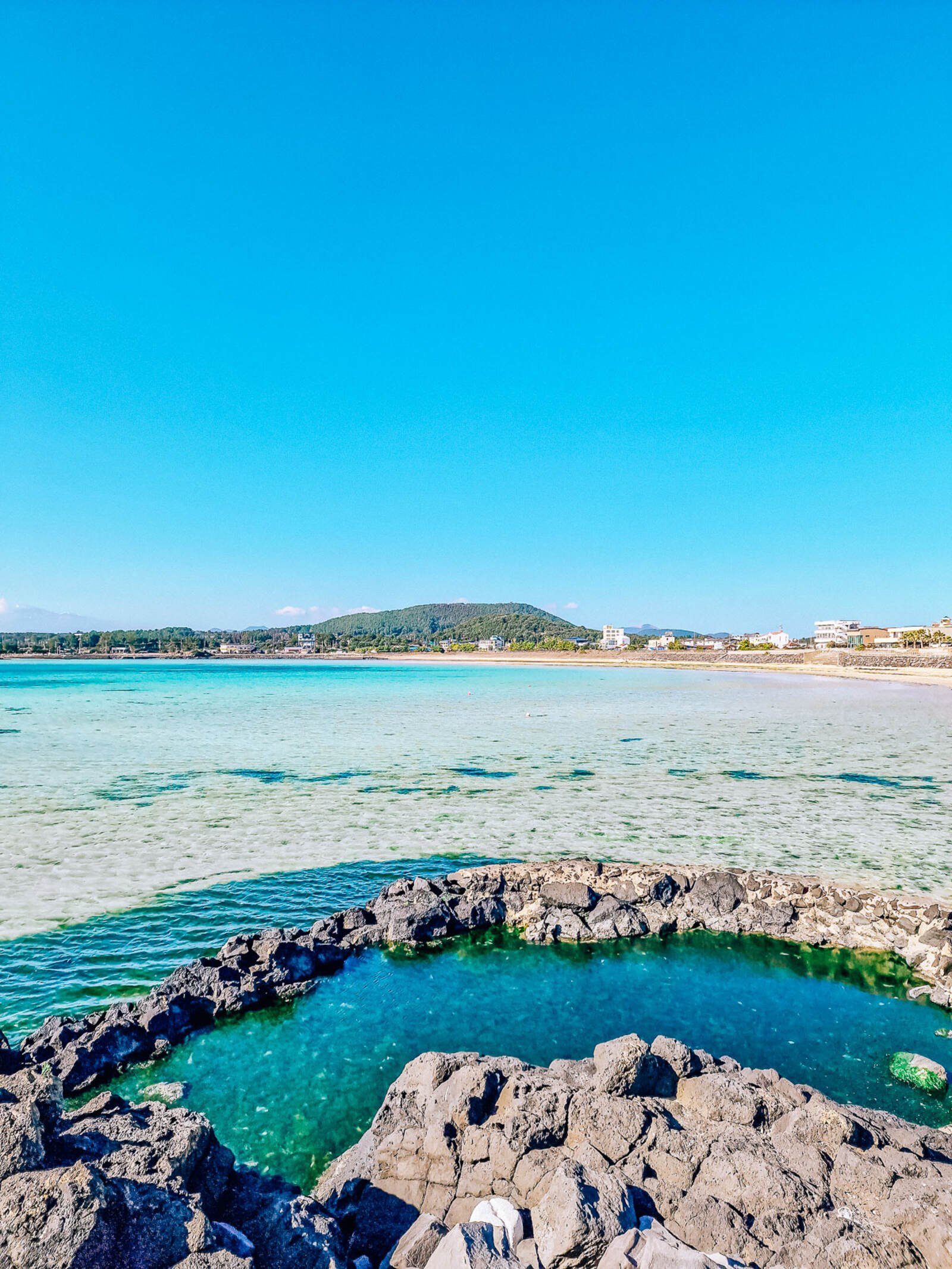 a rock pool surrounded by a calm, turquoise clear bay of water. A beach in the distance on a clear sunny day
