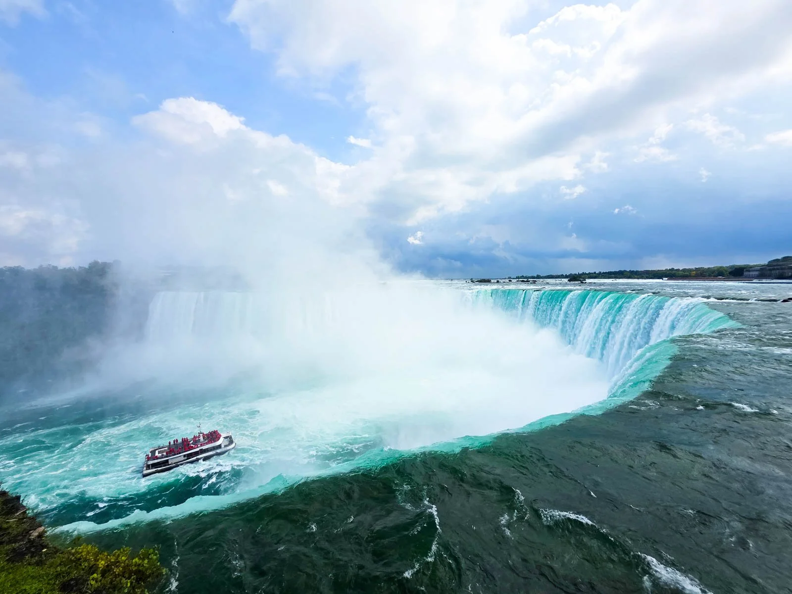A huge horseshoe shaped waterfall with turquoise water flowing over and mist rising from the pool below at Niagara Falls. A cruise ship with people out on deck is seen sailing into the mist