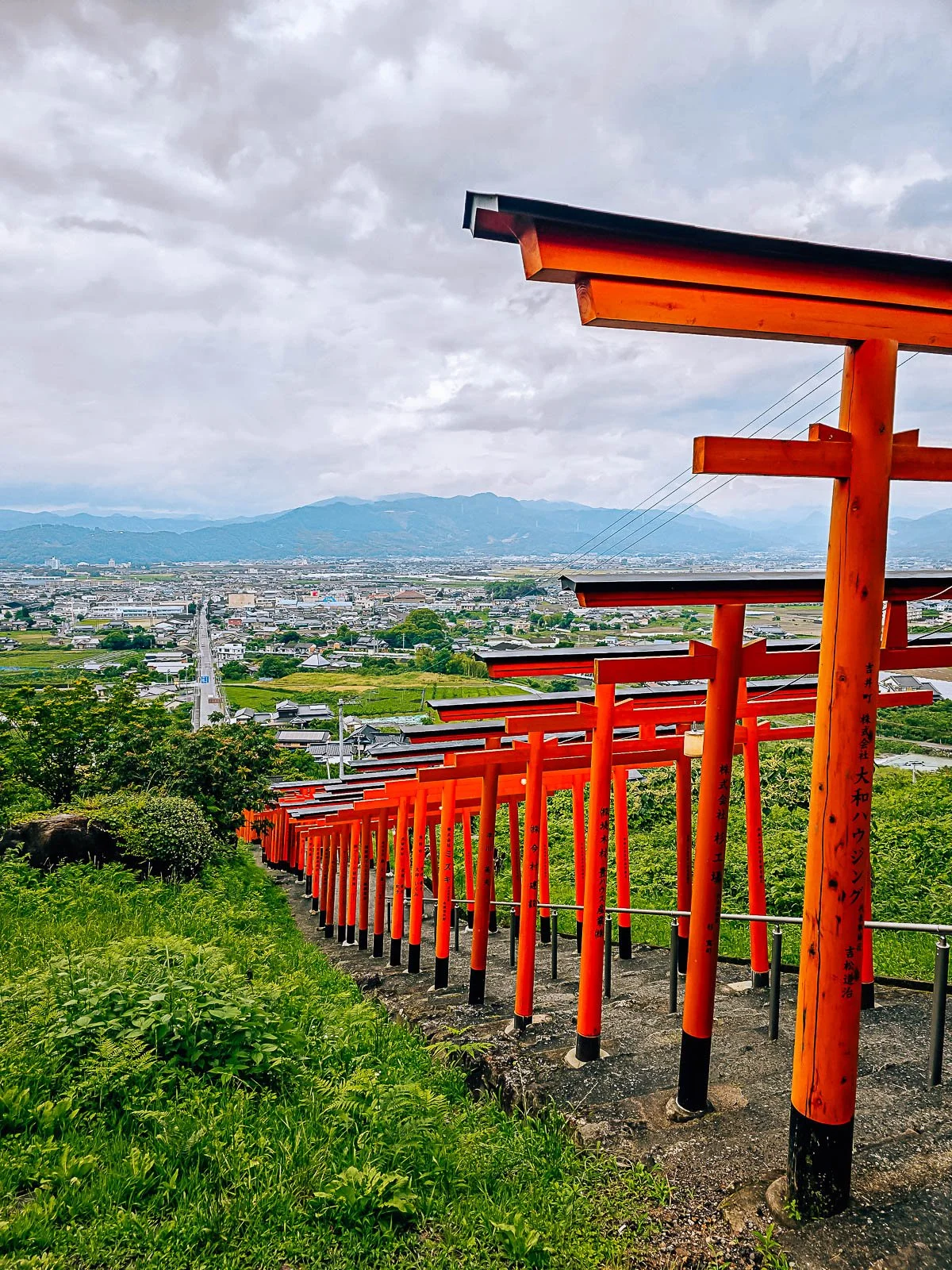 Many orange torii gates in a row over a steps that lead down the side of a hill. The hill is green on both sides of the steps with a town and mountains visible in the distance