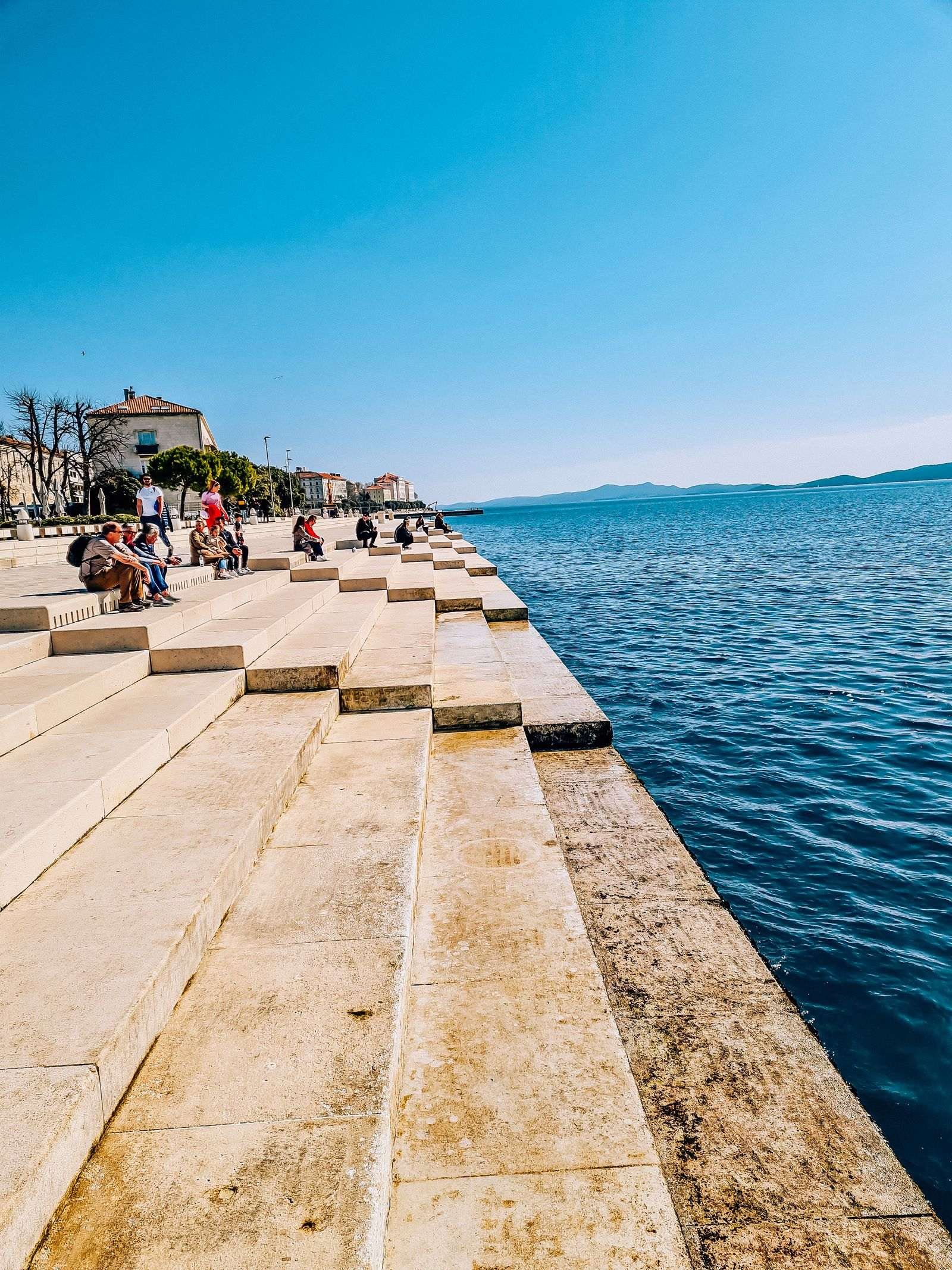 Stone steps leading down to the blue sea with people sitting on the steps in the sun