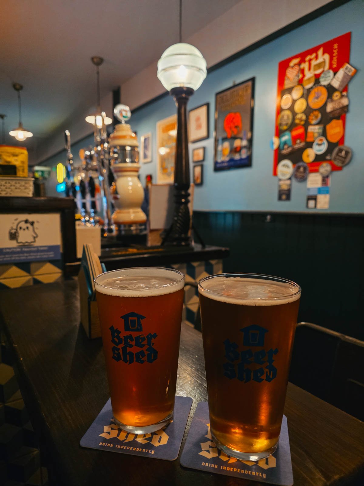 Two beers on a table in glasses that read "beer shed" the bar is visible in the background in a low lit room with lots of pictures on the wall behind