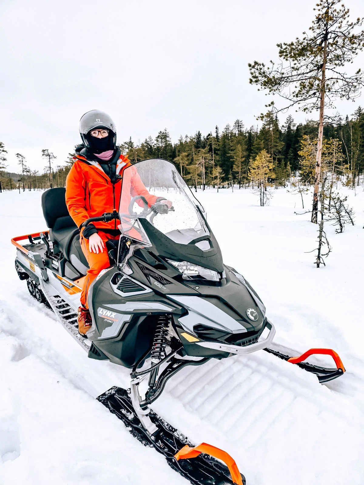 Helena in red driving a large black snowmobile on a snowy trail