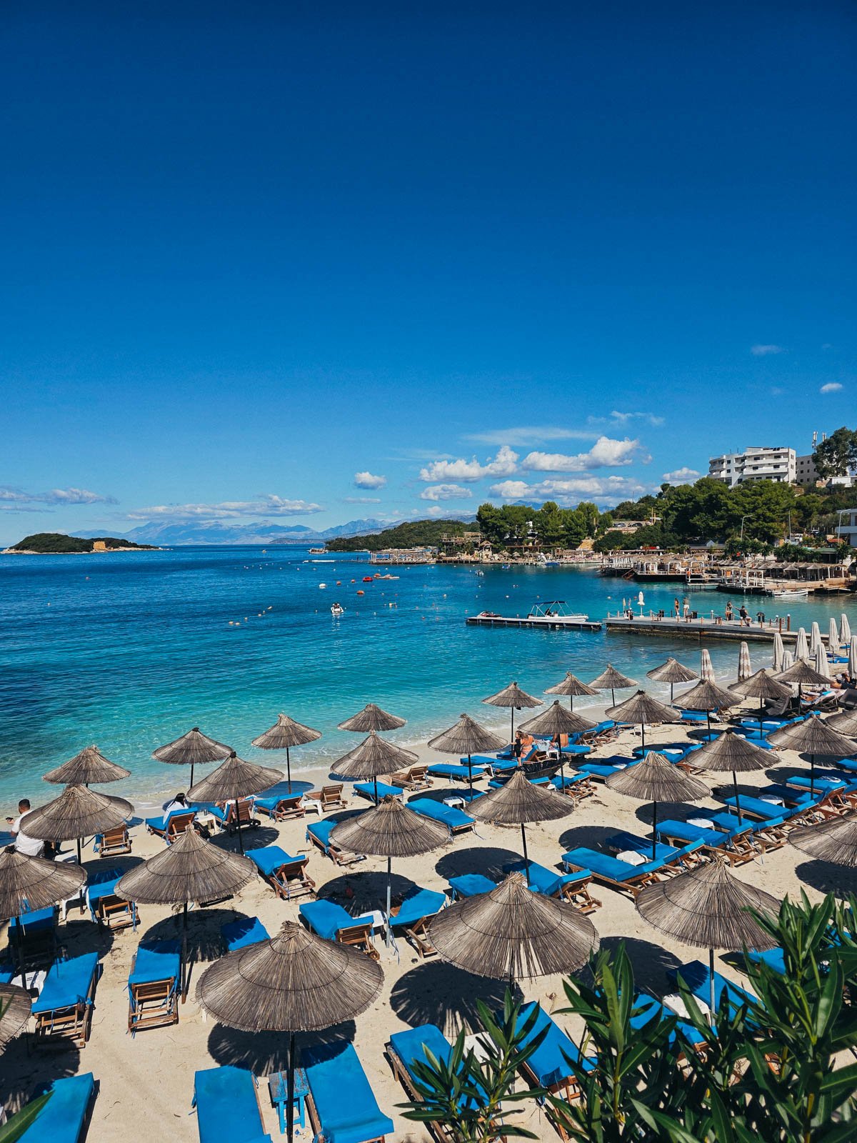 A white sand beach covered in bright blue beach loungers with palm frond umbrellas. The beach is along clear blue water in Ksamil, Albania