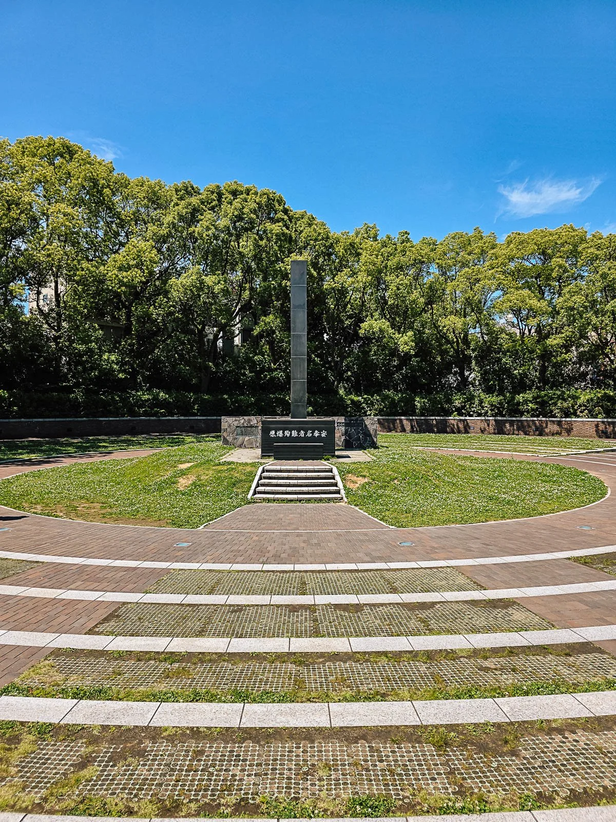 A dark stone monument in a park surrounded by grass and trees. steps lead to the moment and concentric circles surround the monunment on the ground