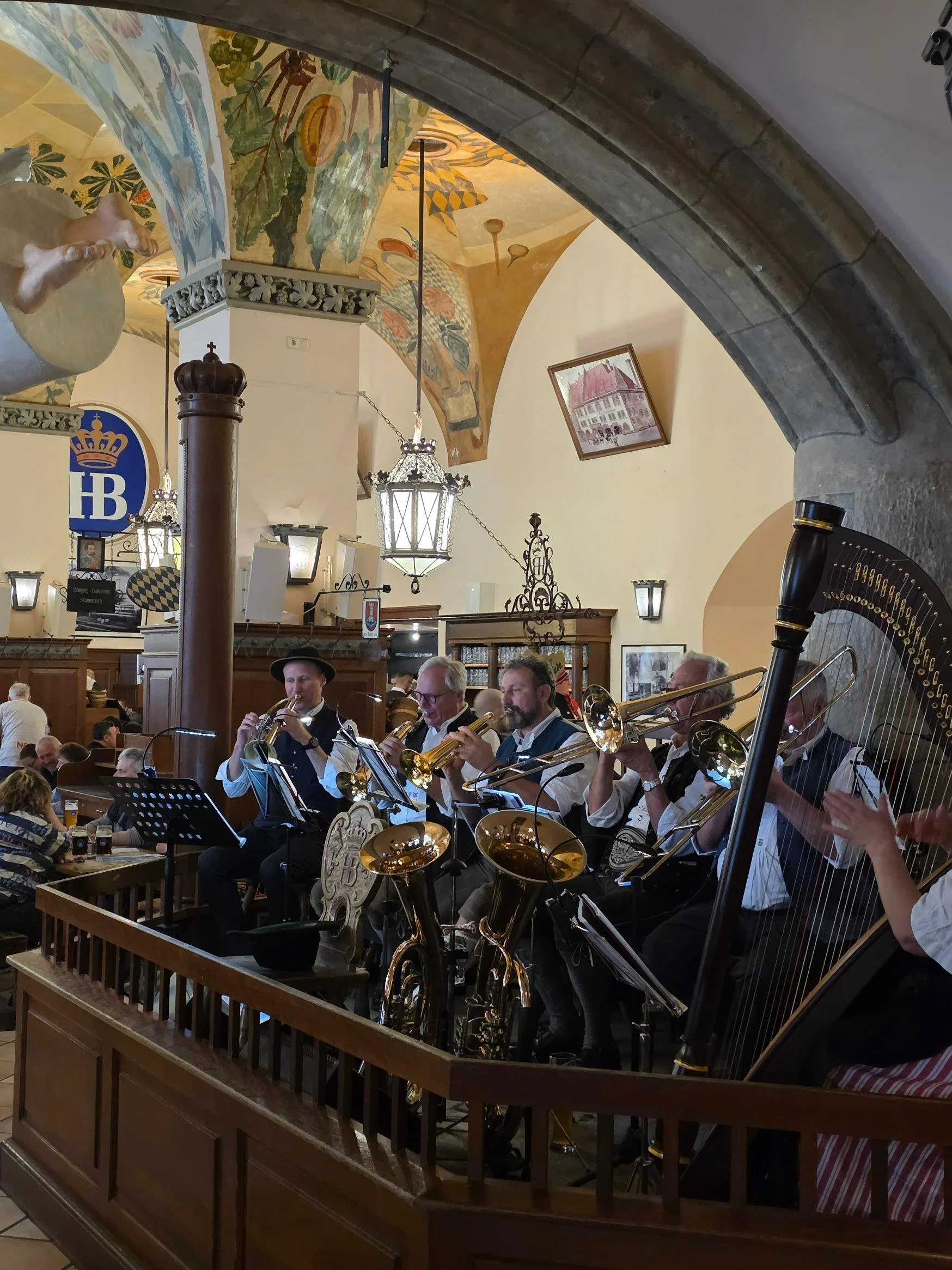 A brass band playing on a small wooden platform in a old large beer hall with murals on the ceiling, and people sitting at tables having beers