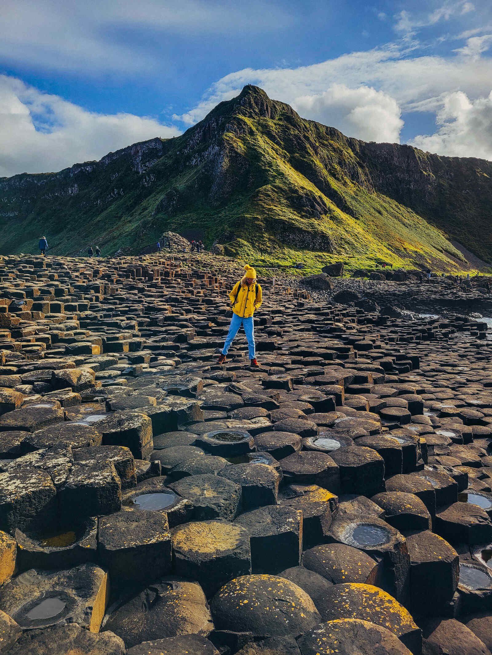 Hundreds of hexagonal rocks on the ground with a girl jumping between them and a green rocky cliff face in the background at the Giant's Causeway in Northern Ireland
