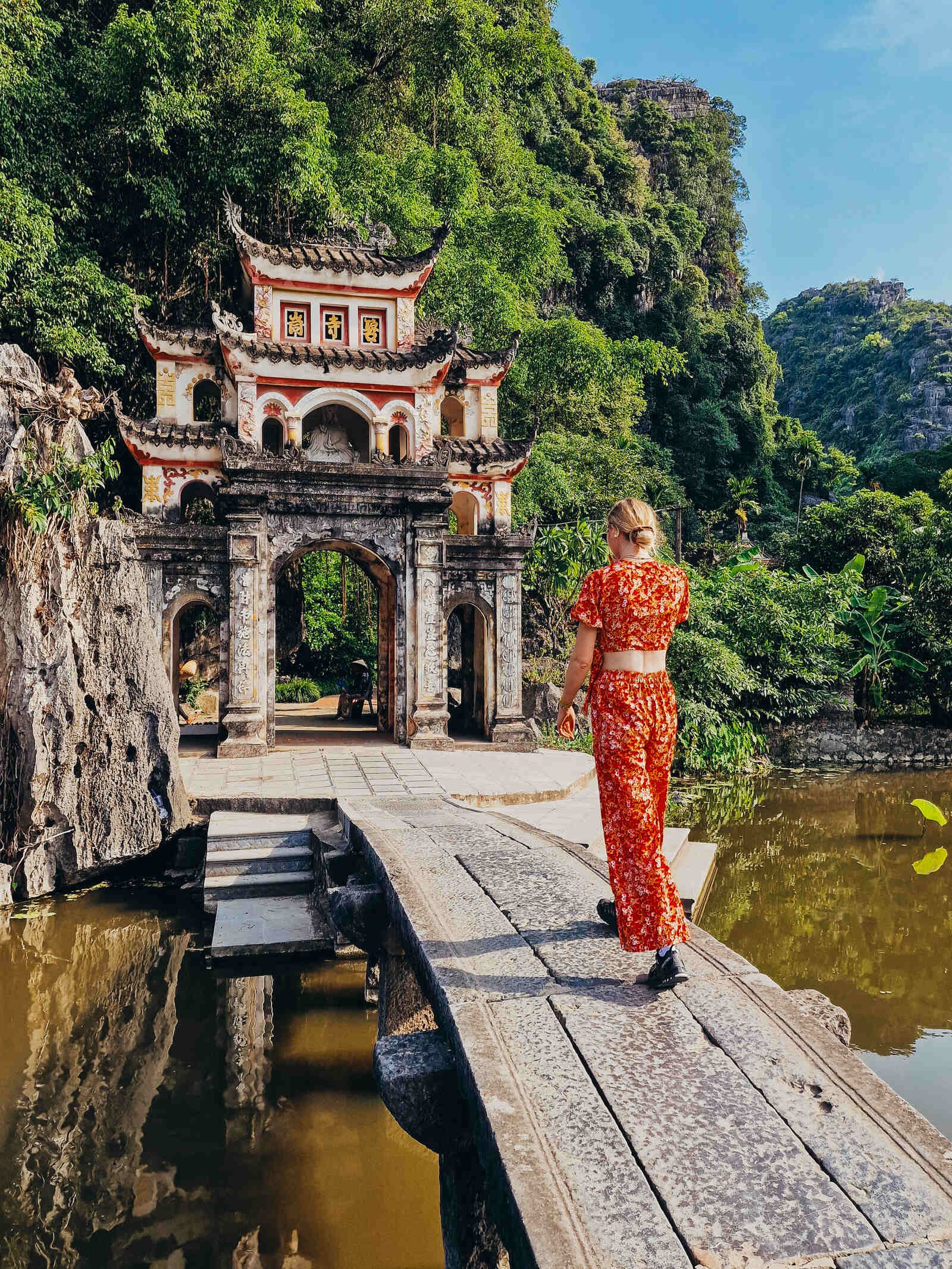 Helena walking across a stone bridge to an ancient pagoda archway surrounded by cliffs