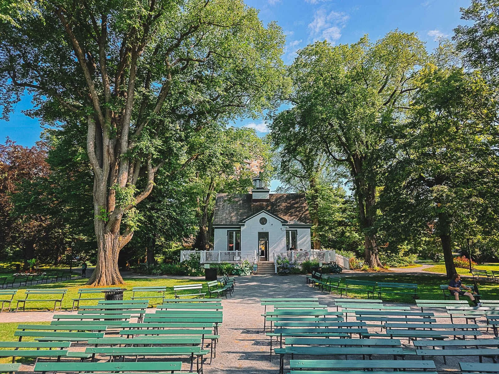A large old white wooden building surrounded by green trees. Many green benches are placed in front of the building