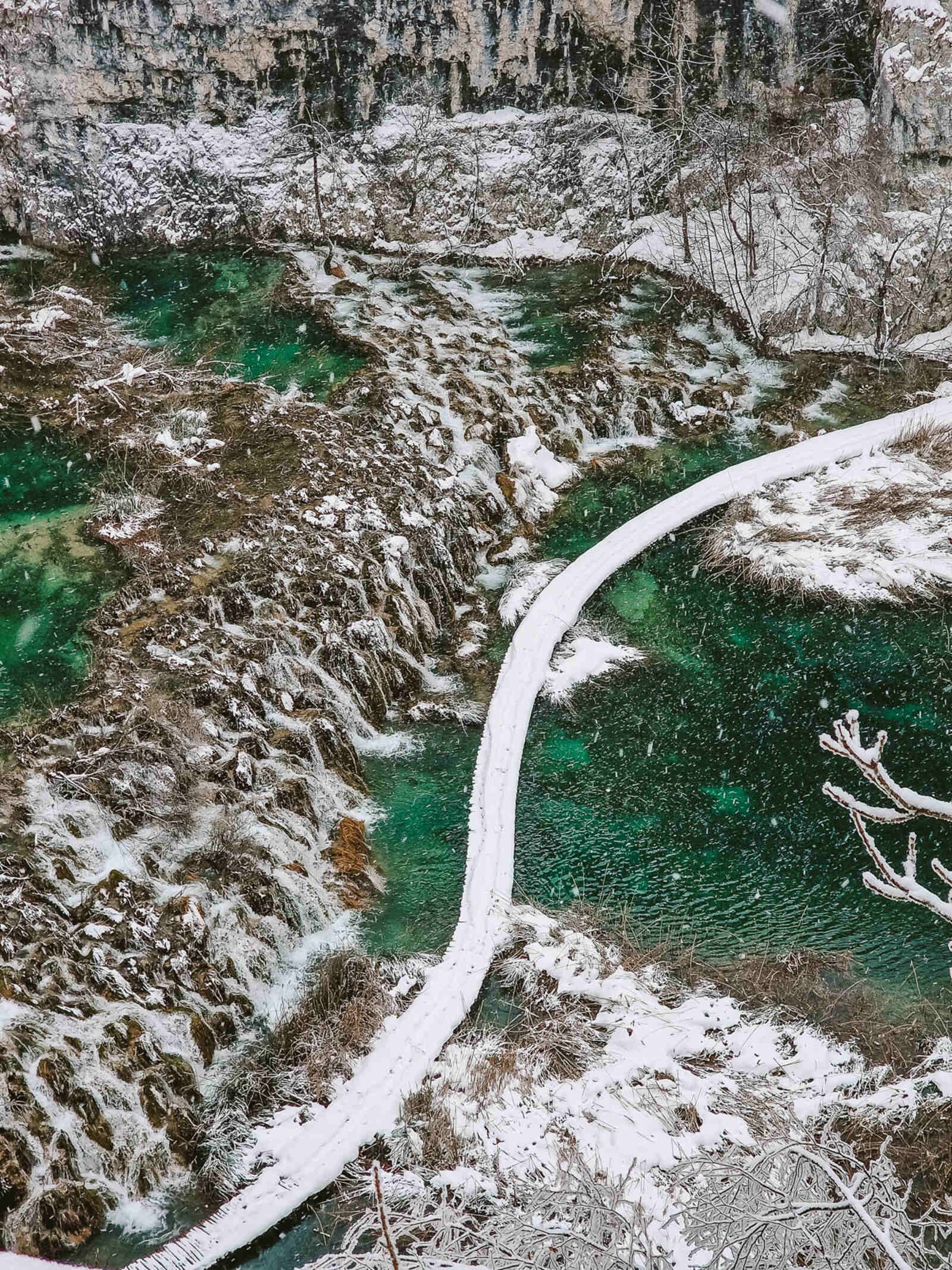 Looking down a cliff to a turquoise blue green lake below with small waterfalls falling into it and a white, snow covered pathway winding above the water
