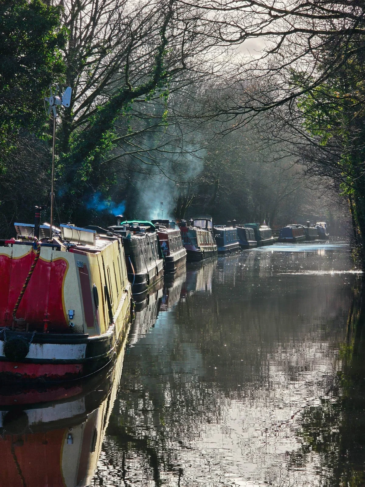 10 canal boats lined along the left side of the canal, smoke rises from several of them