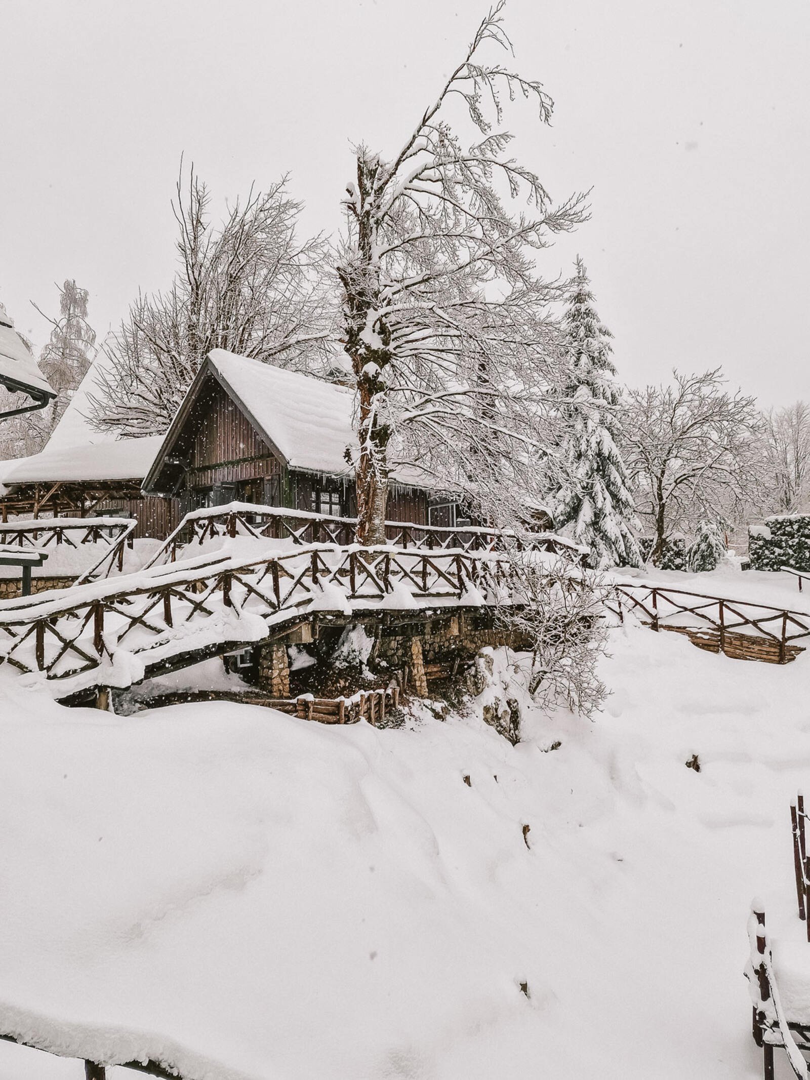 Plitvice hotel ethno houses, wooden cabin style building with a wooden fence running alongside with deep snow around it, snow covered roof, snow covered trees and snow falling in the photo