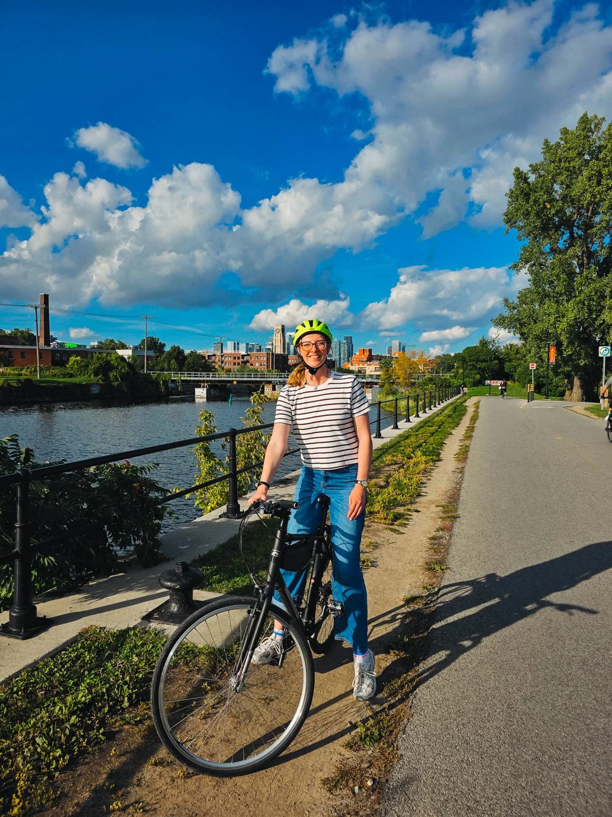 Helena stopped on the side of a bath on a bike, smiling at the camera, wearing a neon yellow helmet with the city skyline behind her