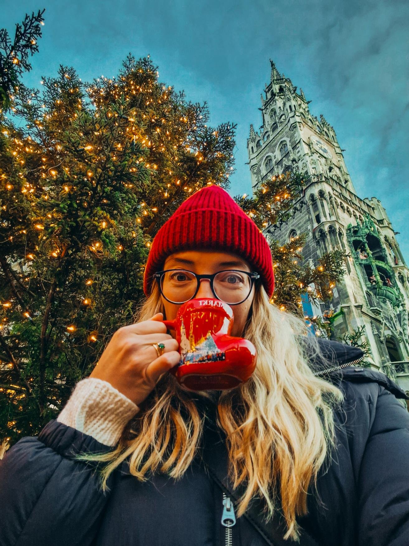 Helena drinking from a bright red mug with a large lit up christmas tree and tall ornate stone tower behind her