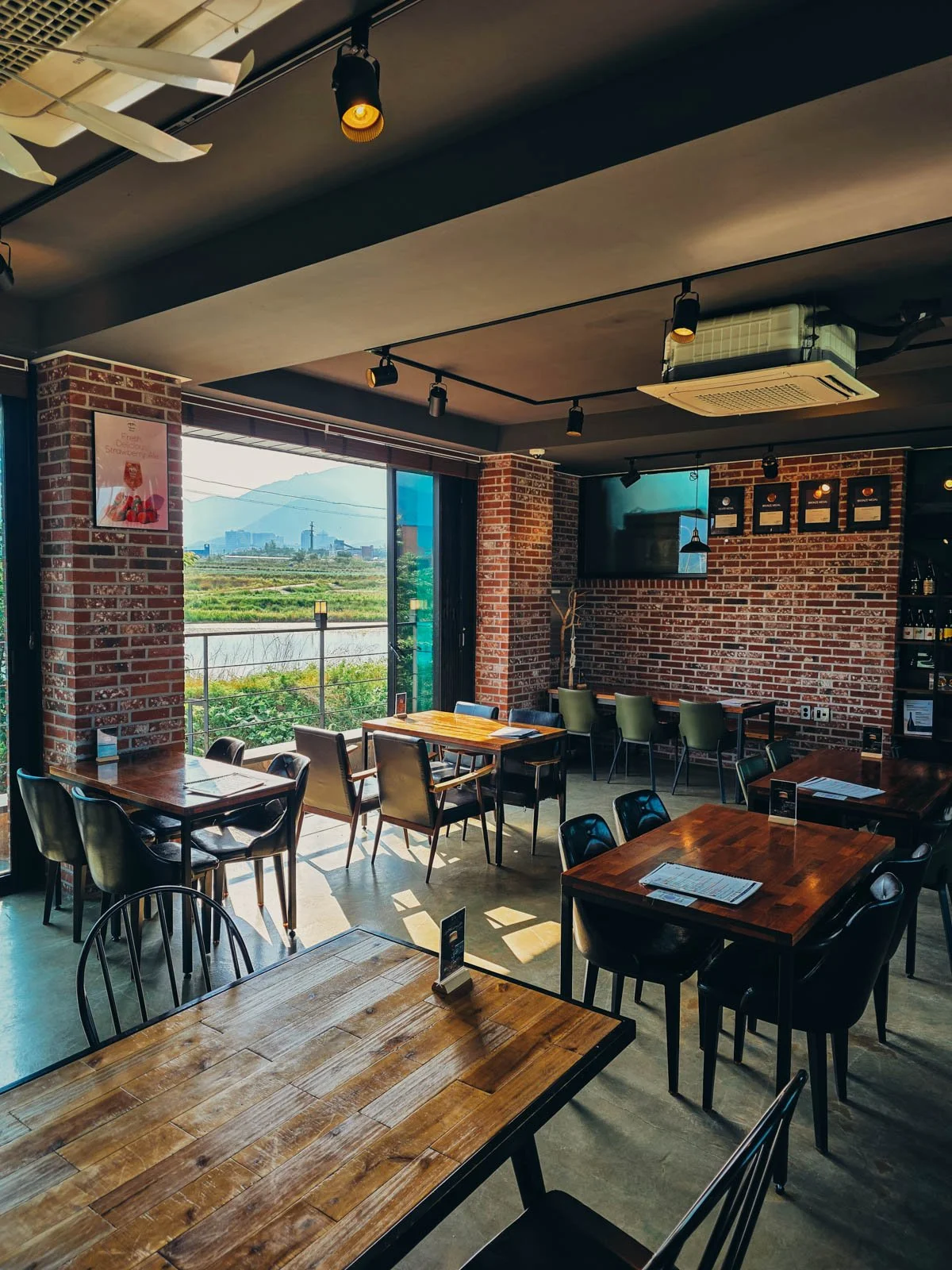 An industrial style bar with exposed brick and 6 tables of different sizes with a door to a balcony open on the left. The view out the door is of fields and mountains beyond