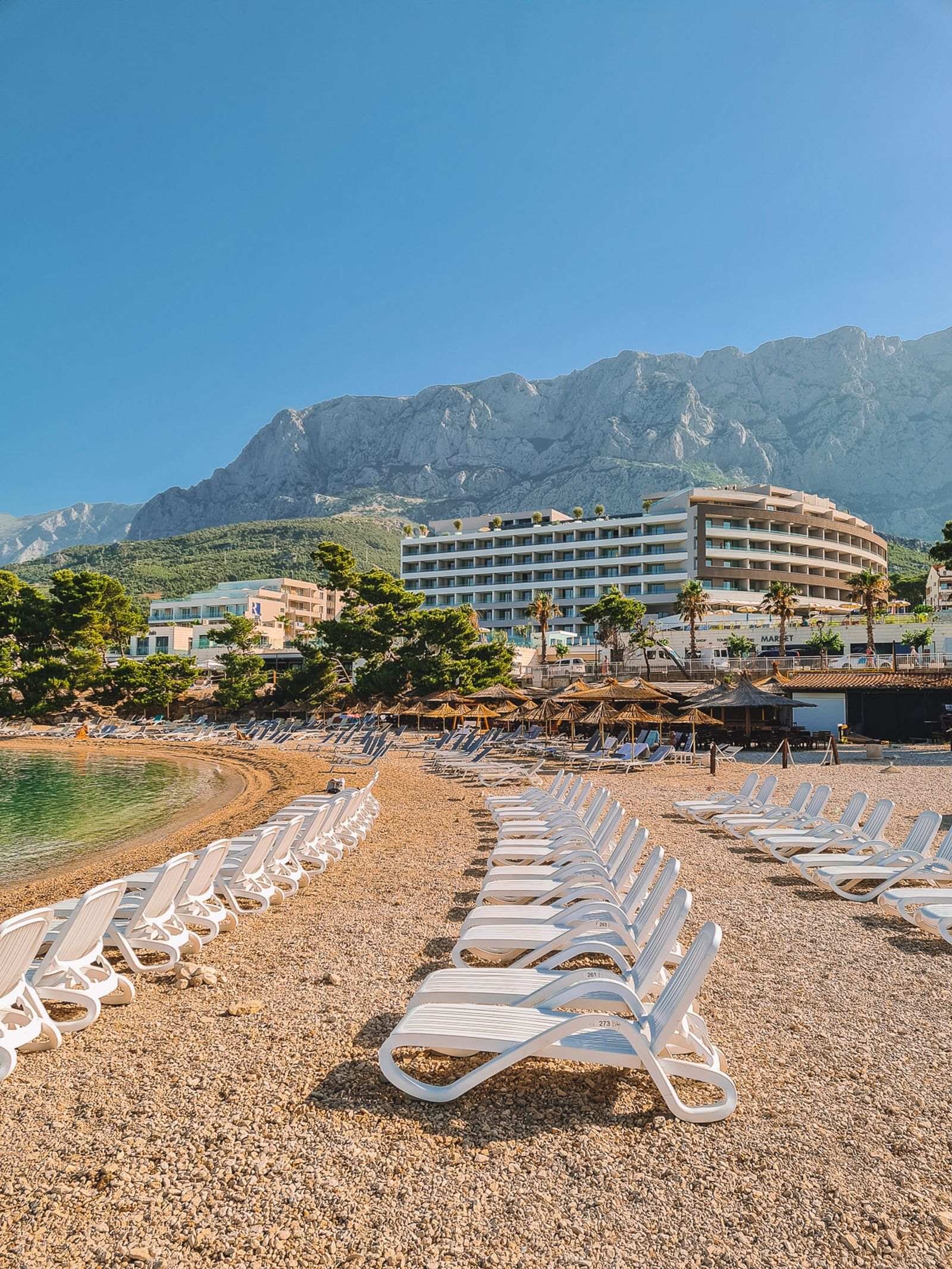 many white sun loungers laid out on the beach with hotels in the background and mountains towering over the town
