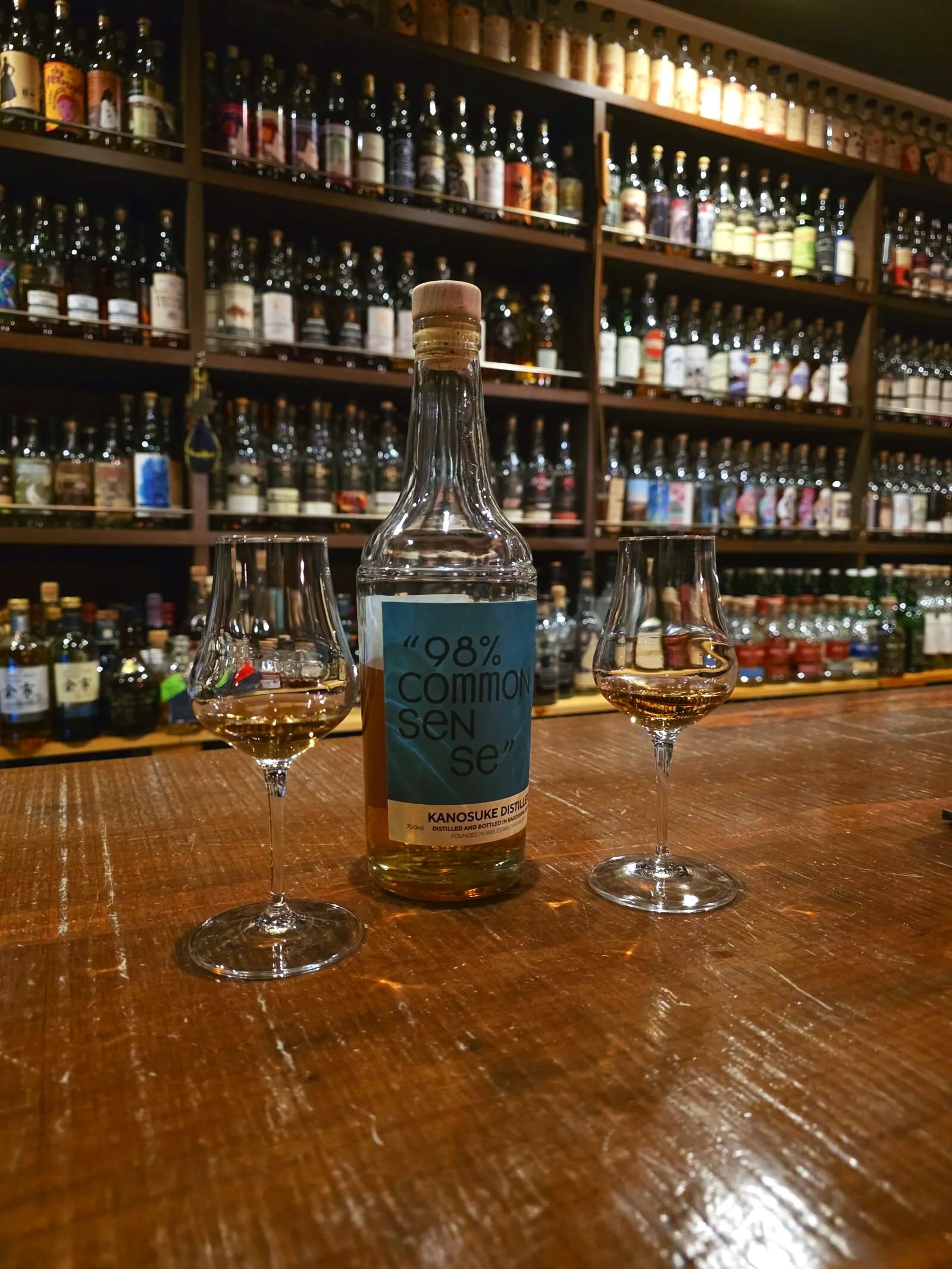 two small glasses either side of a whisky bottle on a wooden bar surface and shelves behind with hundreds of bottles of whisky lined up on them