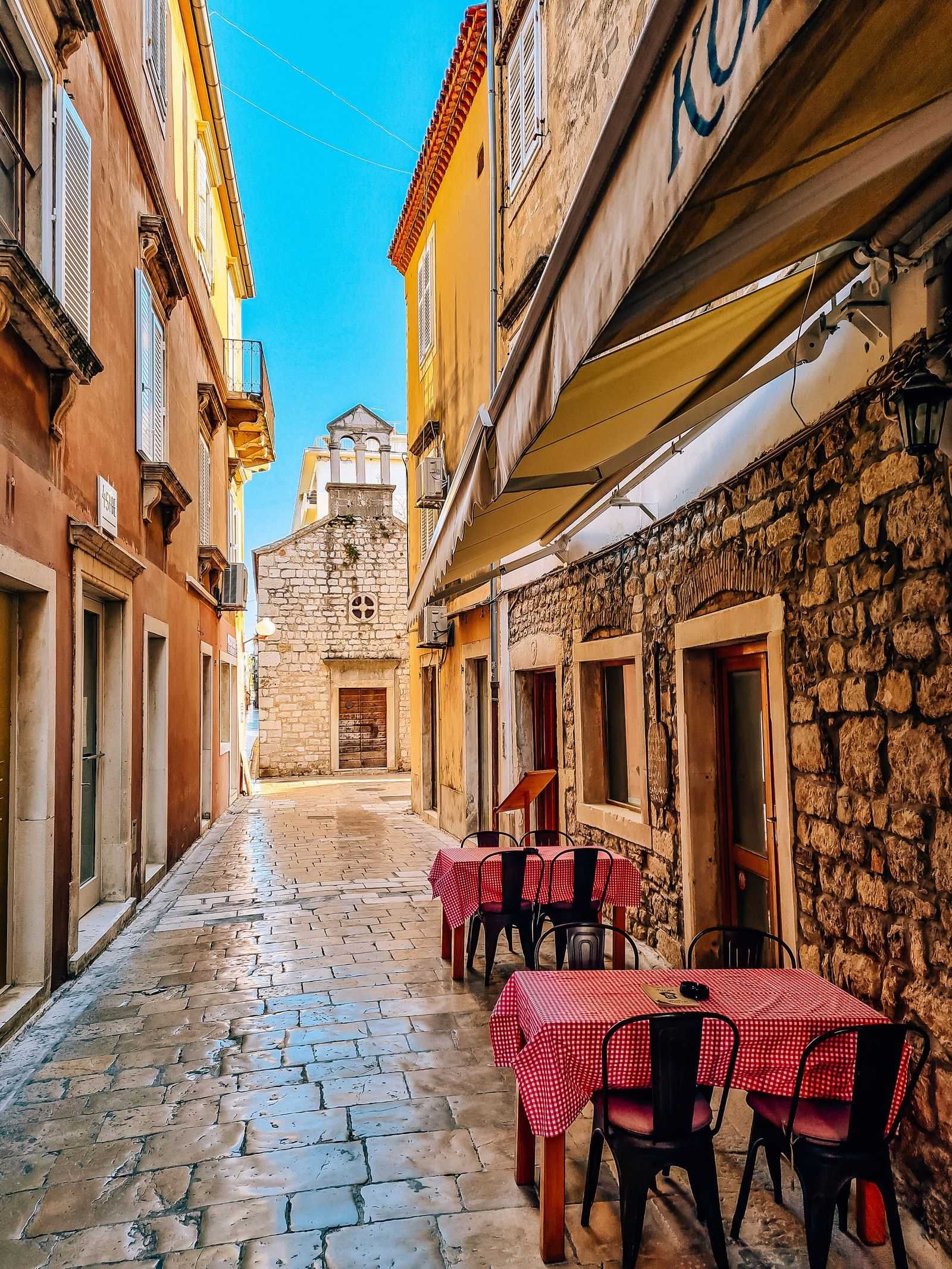 Red checked table clothed outdoor seating outside a stone restaurant with a narrow polished stone street leading to a old church.