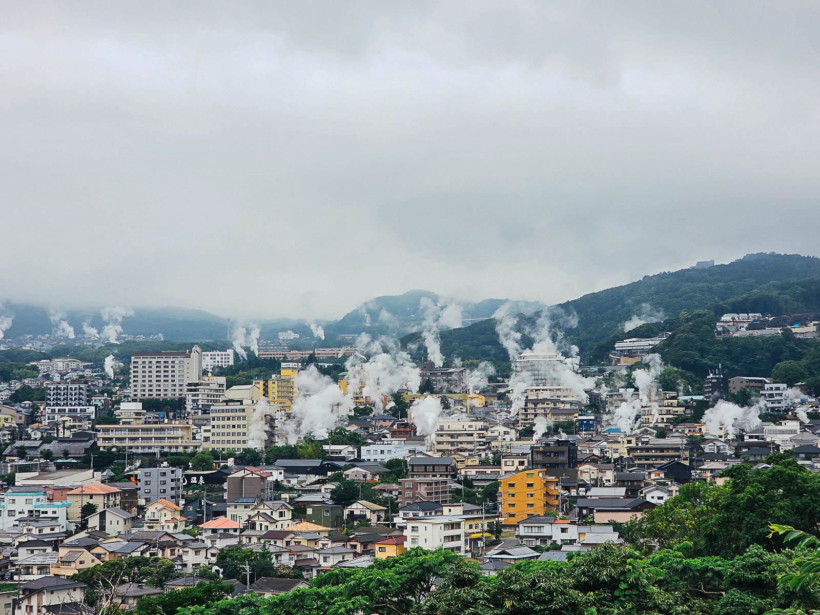 A city scape of Beppu hot spring town surrounded by green hills and many plumes of steam rising all over the city from the hot springs
