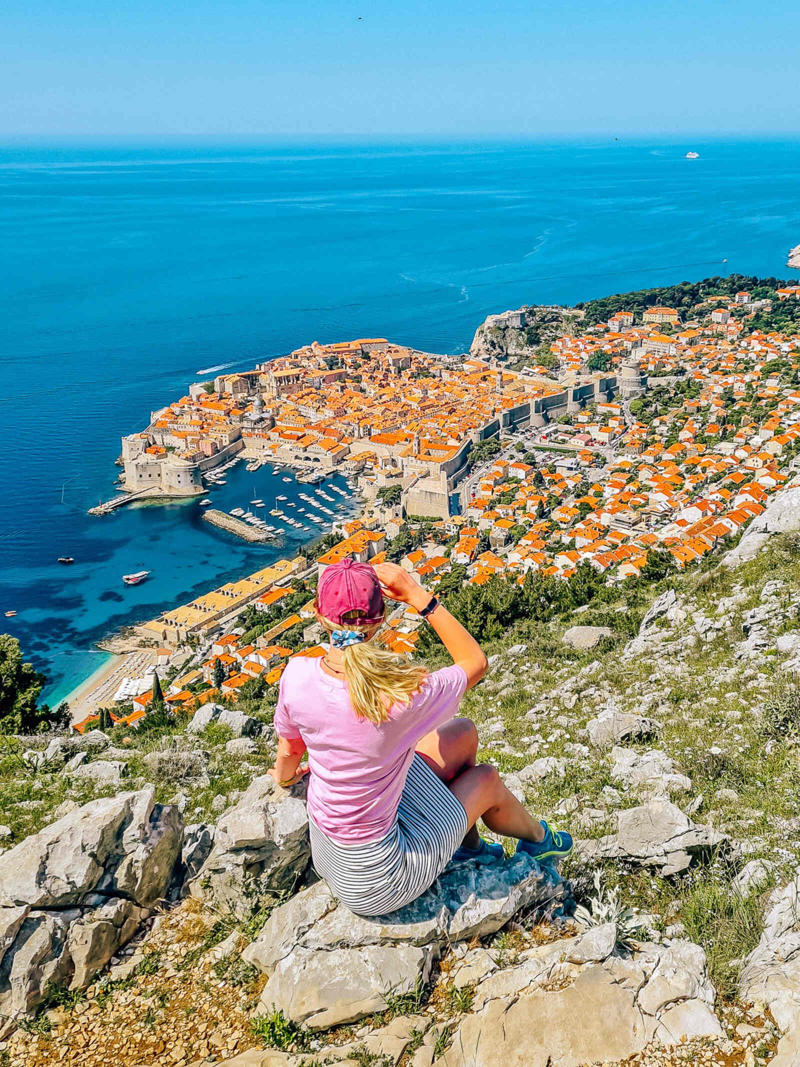 Girl in a pink shirt and hat with a stripe skirt sitting on a rock on a mountain side and looking down on the historic walled town of Dubrovnik, surrounded by blue sea