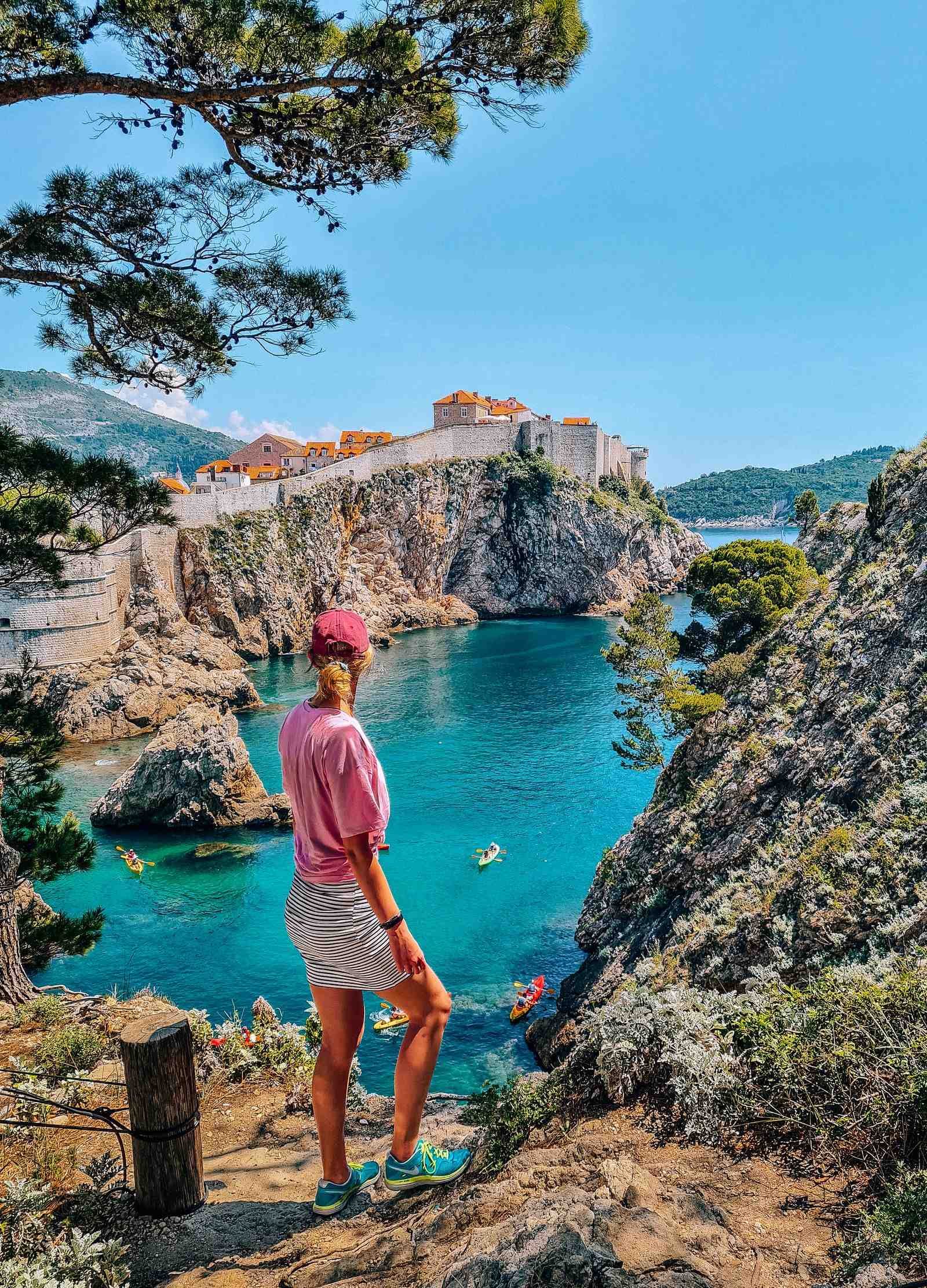 A girl in a pink shirt standing alongside a cliff with blue water and the walled old town of Dubrovnik along a cliff in the distance