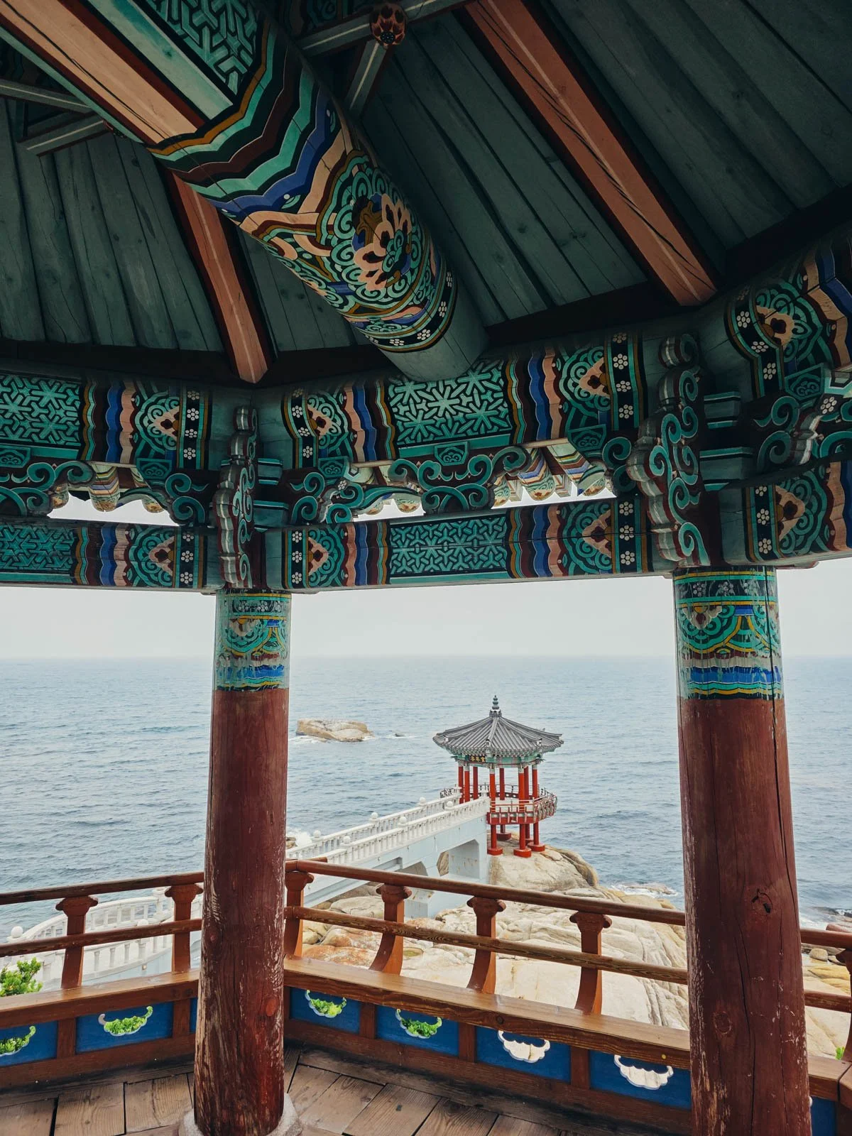 Standing under a wooden pavillion with red pillars and a balcony with overlooks the sea where a rocky outcrop is reached by a white walkway. At the end of the walkway is another temple pavilion with red pillars and a green and blue roof and patterns