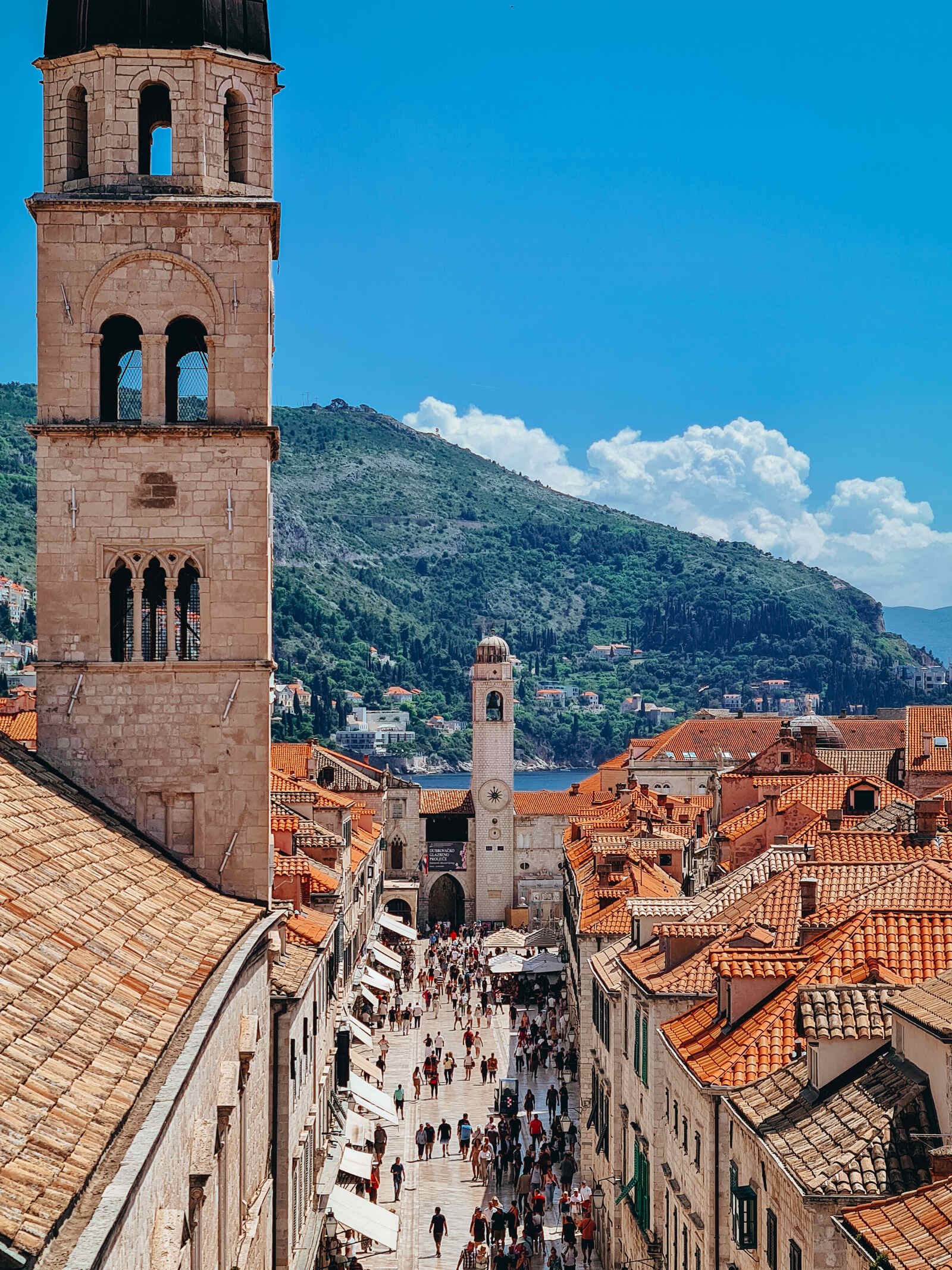 A view from the Dubrovnik city walls looking down the famous Stradun Street in Dubrovnik Old Town. The street is full of people walking and lined with stone houses and orange rooftops. There is a church bell tower in the foreground and at the bottom