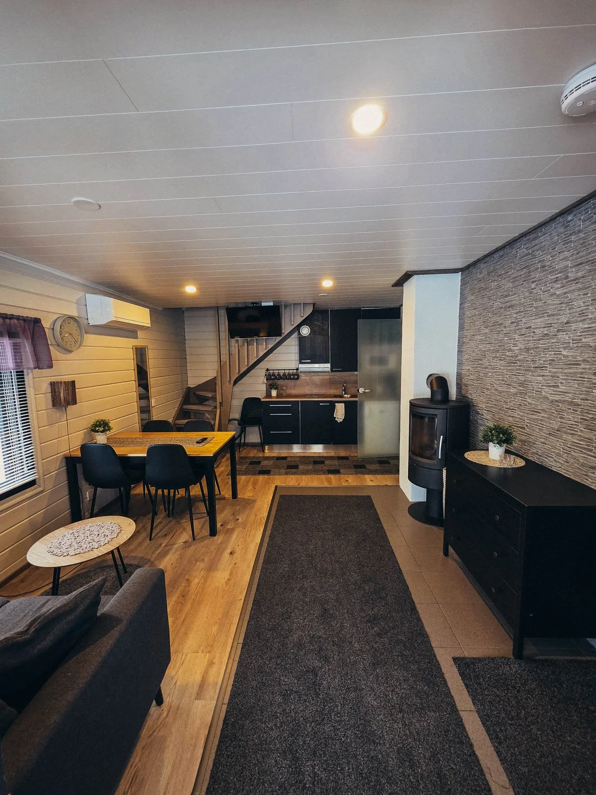 The cabin interior with a wood floor long rug, wood table with chairs, kitchen, a black log burner, and stairs going up to the second floor in the back of the cabin.