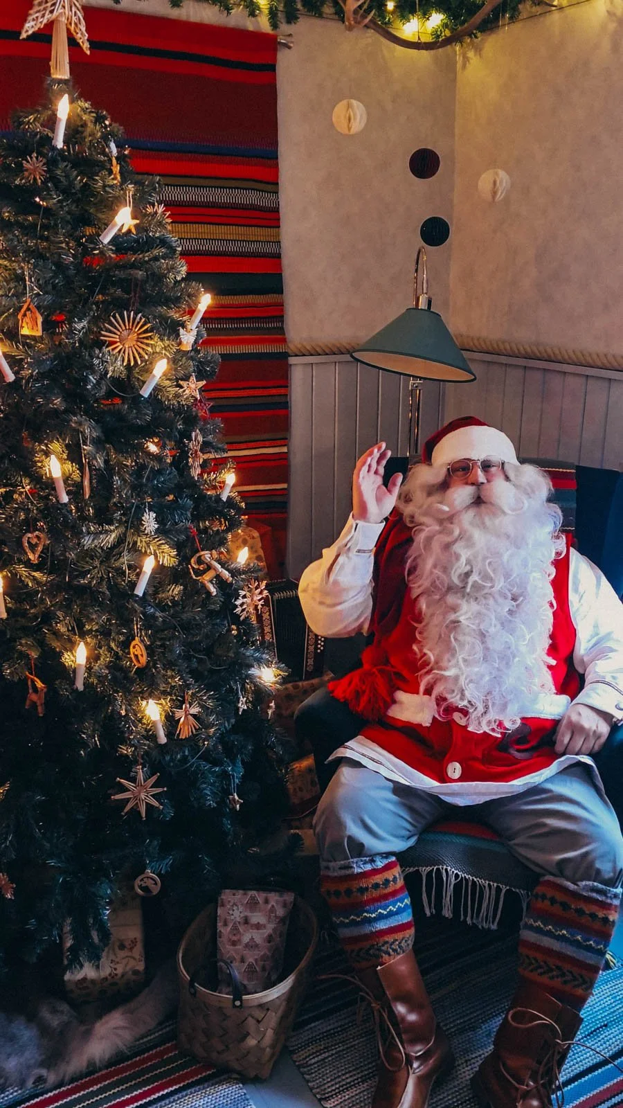 Santa with a big white beard and red outfit sitting in a green chair next to a lit up Christmas tree