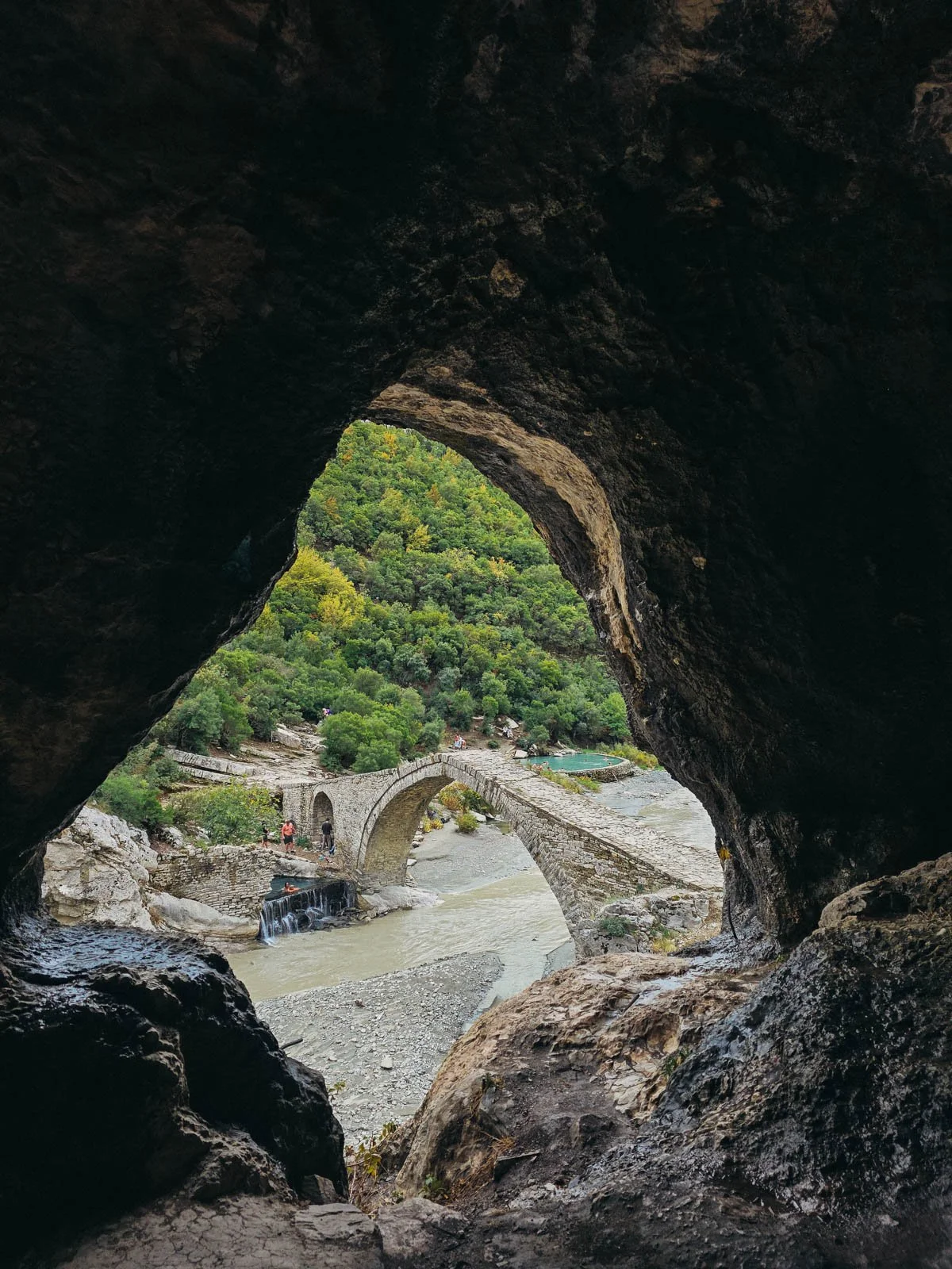 Looking out of a cave opening with a large entrance that looks out at a stone bridge crossing a river