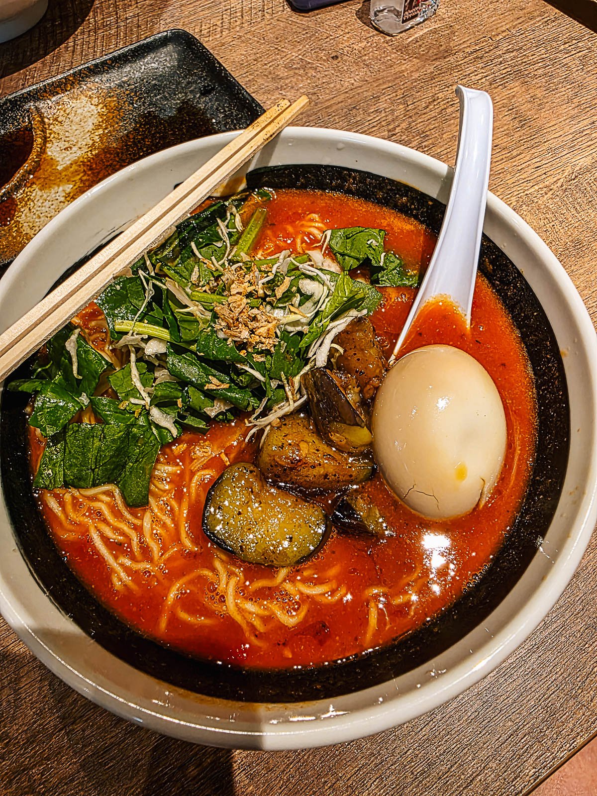 A bowl of ramen in a white bowl with a boiled egg, aubergine, noodles and greens, The broth is red because it's tomato based broth
