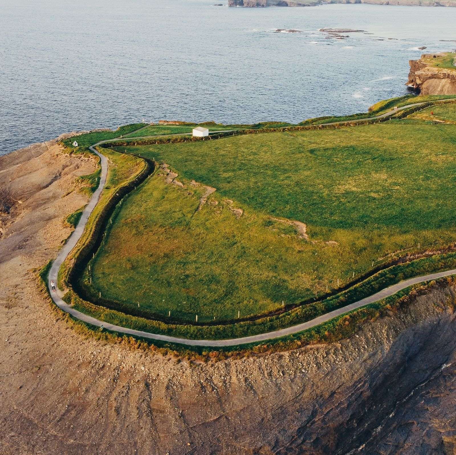 drone photo of a walking path following the edge of a grassy cliff with rockface leading to the water