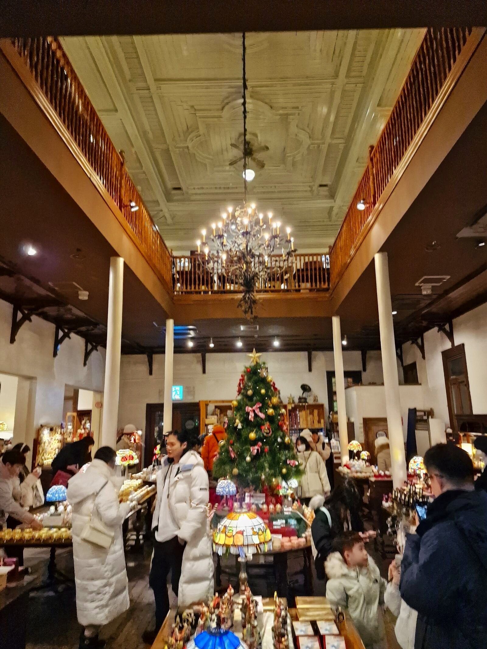 a large old room with vaulted ceiling and a chandelier, a christmas tree in the middle of the room and a few people walking around tables with displays on