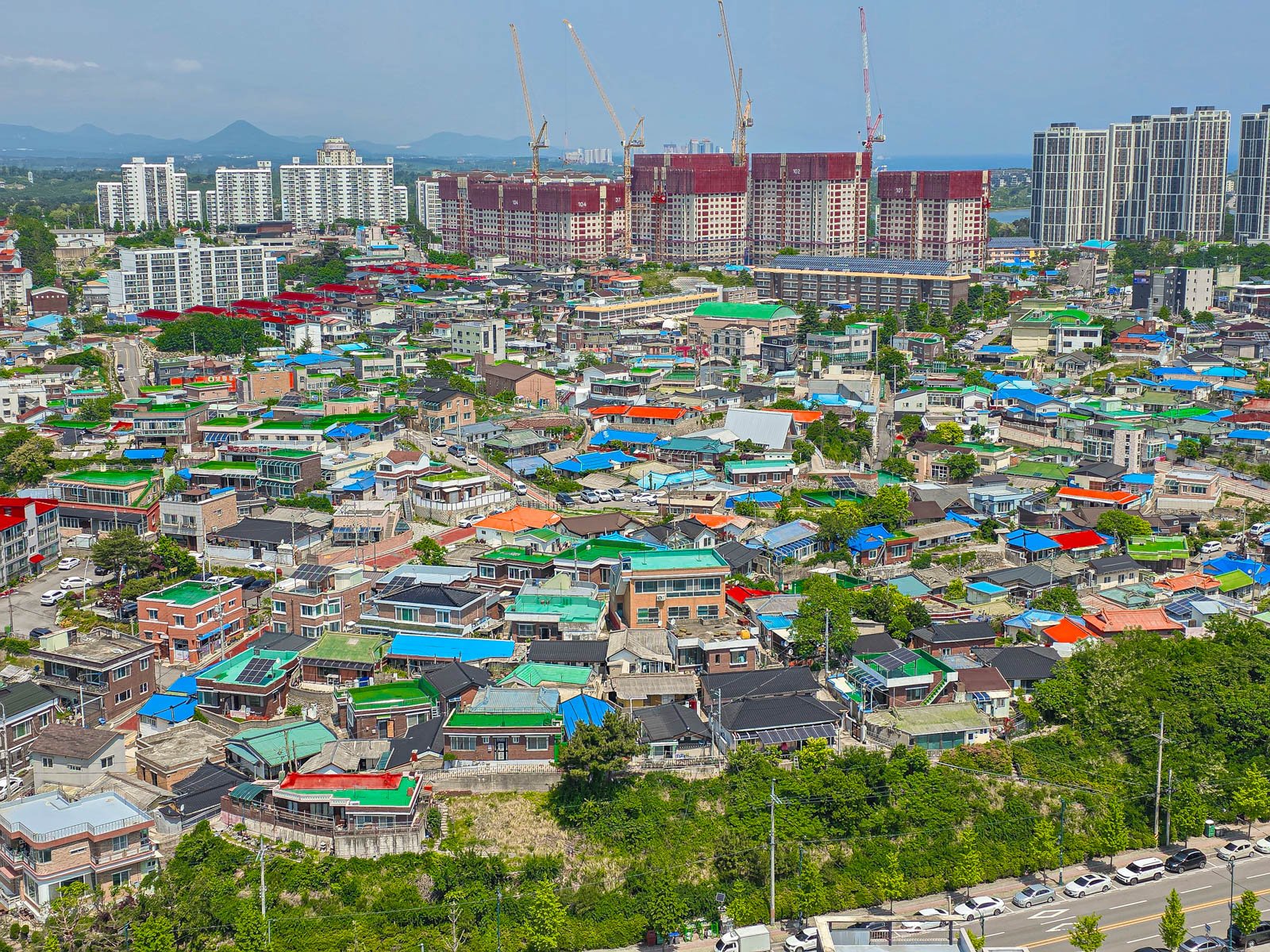 A view looking down on streets with many small buildings with colourful rooftops: red, green, blue, orange and a city skyline with cranes in the distance