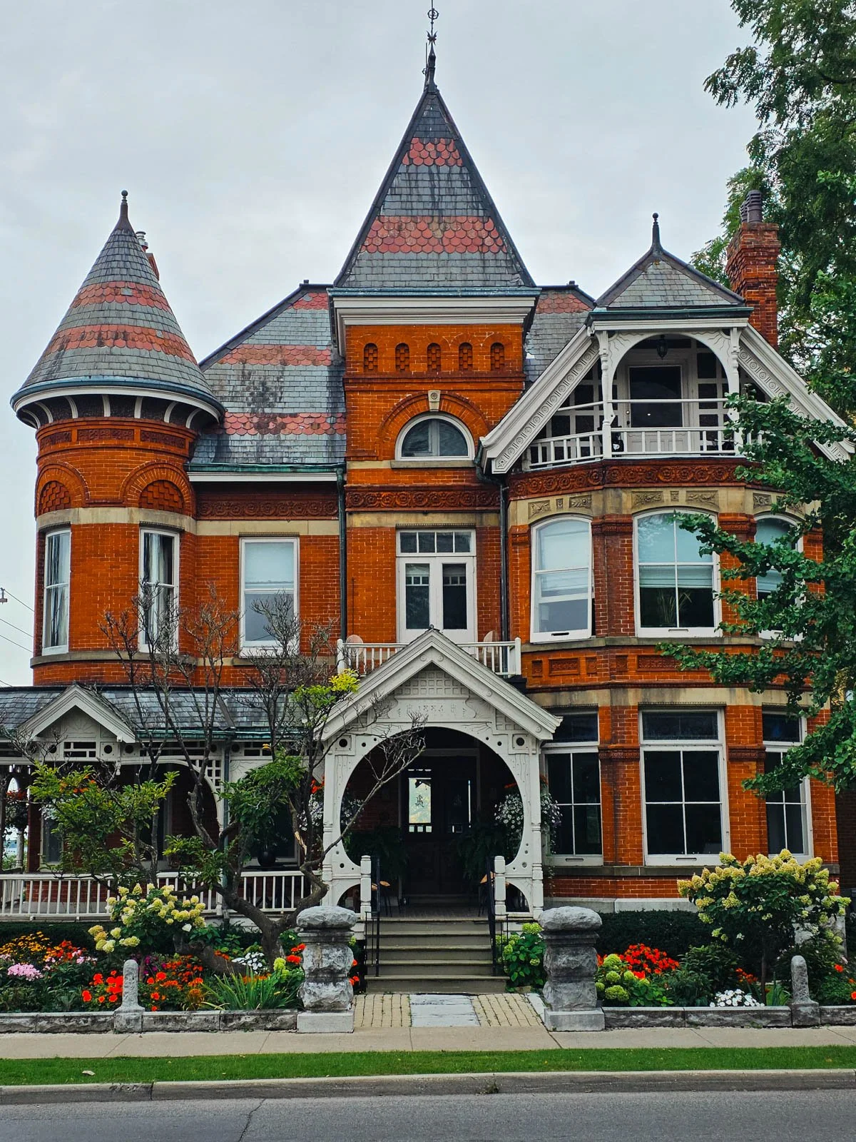 A large, ornate red brick house with many windows, brick turrets ,and a wraparound white porch. The garden at the front is full of colourful flowers