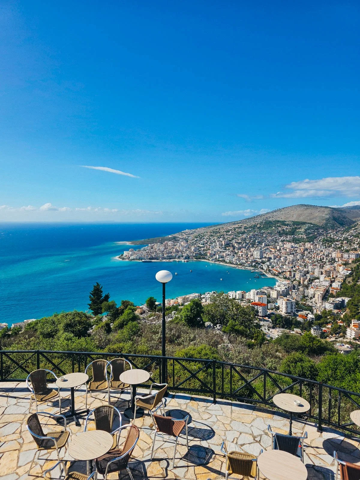 An outdoor seating area on a stone balcony overlooking a costal town with many buildings along clear blue water