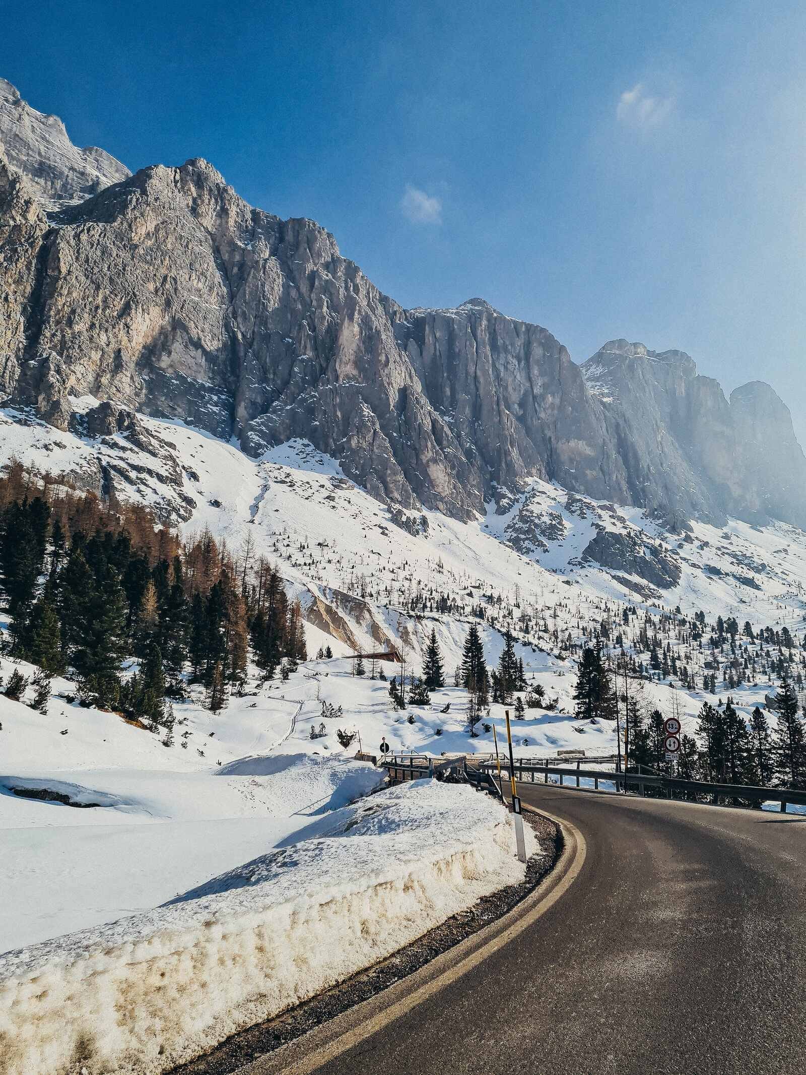 a clear road with snow banks either side and blue sky with jagged grey rocks towering into the sky in Dolomites italy