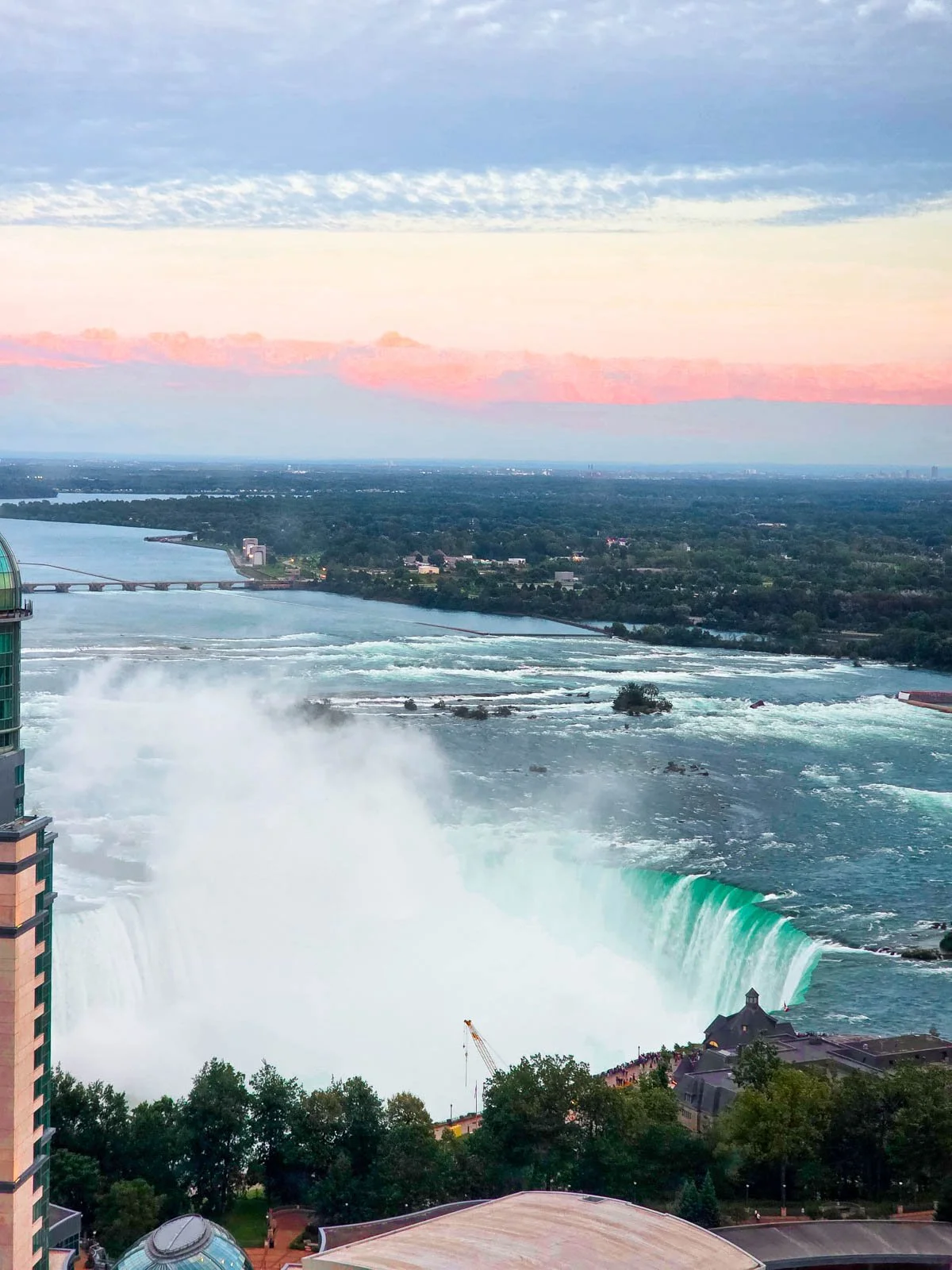 An aerial view of Niagara Falls with the mist rising from the pool and Niagara River visible behind, the sky is pink at sunset