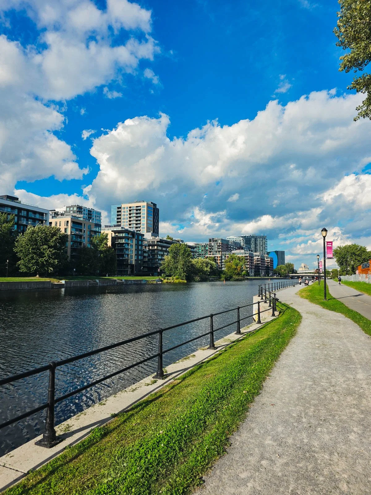 A path runs alongside a canal with a city skyline visible beyond on a blue sky sunny day