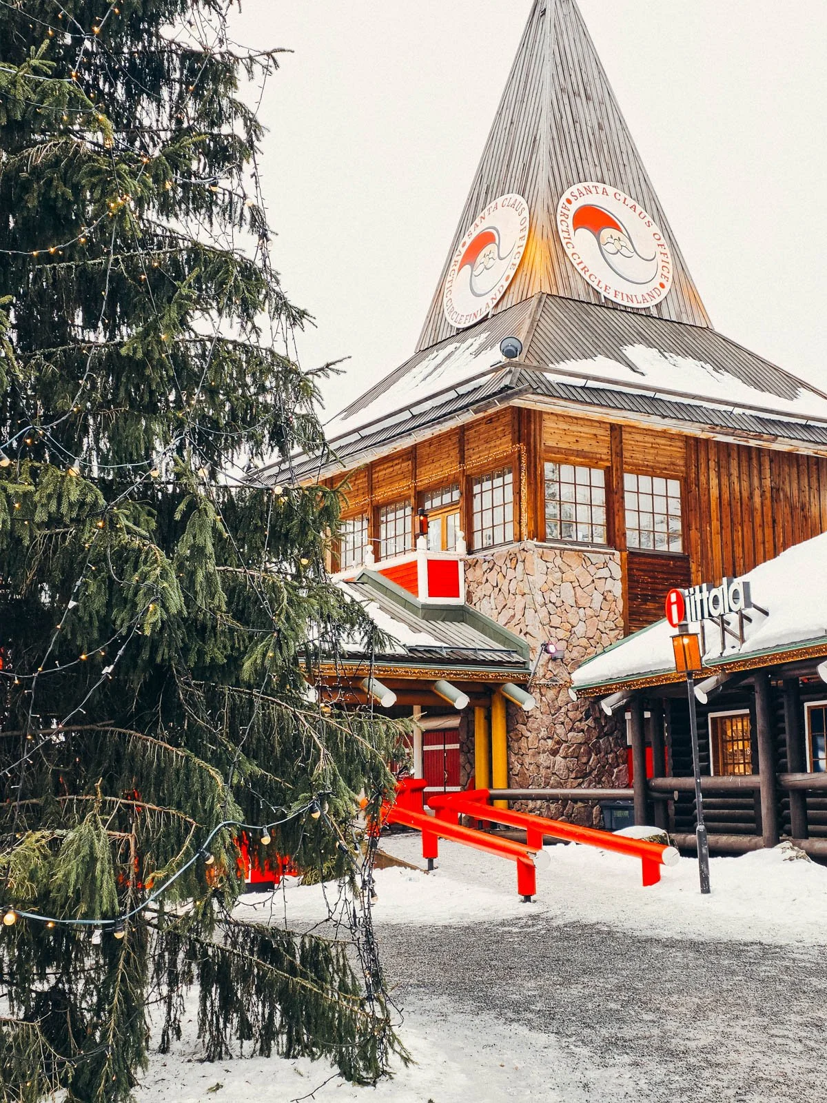A large wooden building with a pointed roof surrounded by snow. A large green ping tree is in front of the building at Santa Claus Village - a must visit on a Finland winter itinerary