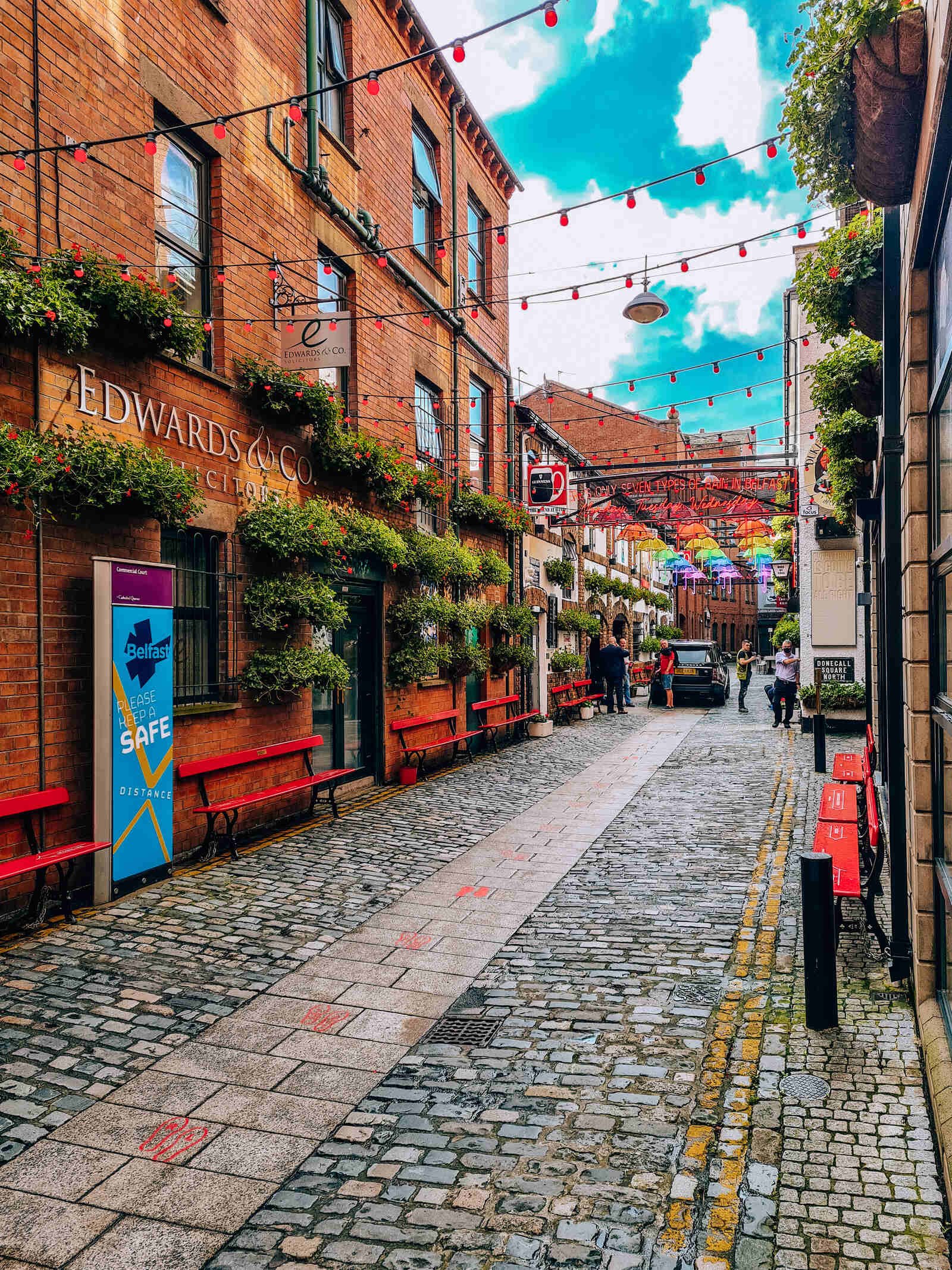 A cobbled street lined with red brick buildings and colourful lights and bunting hanging overhead in Belfast's cathedral quarter on a short break weekend in the UK