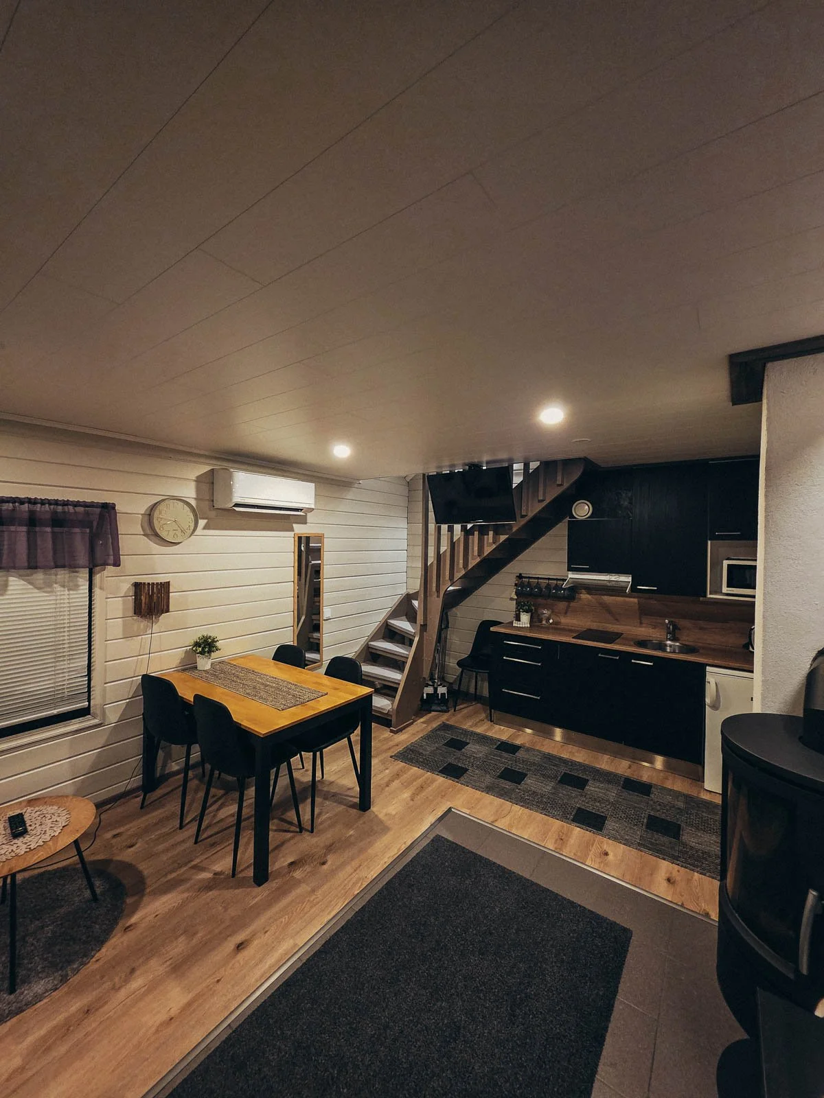 A cabin interior with a wood floor, wood table, kitchen, and a black log burner