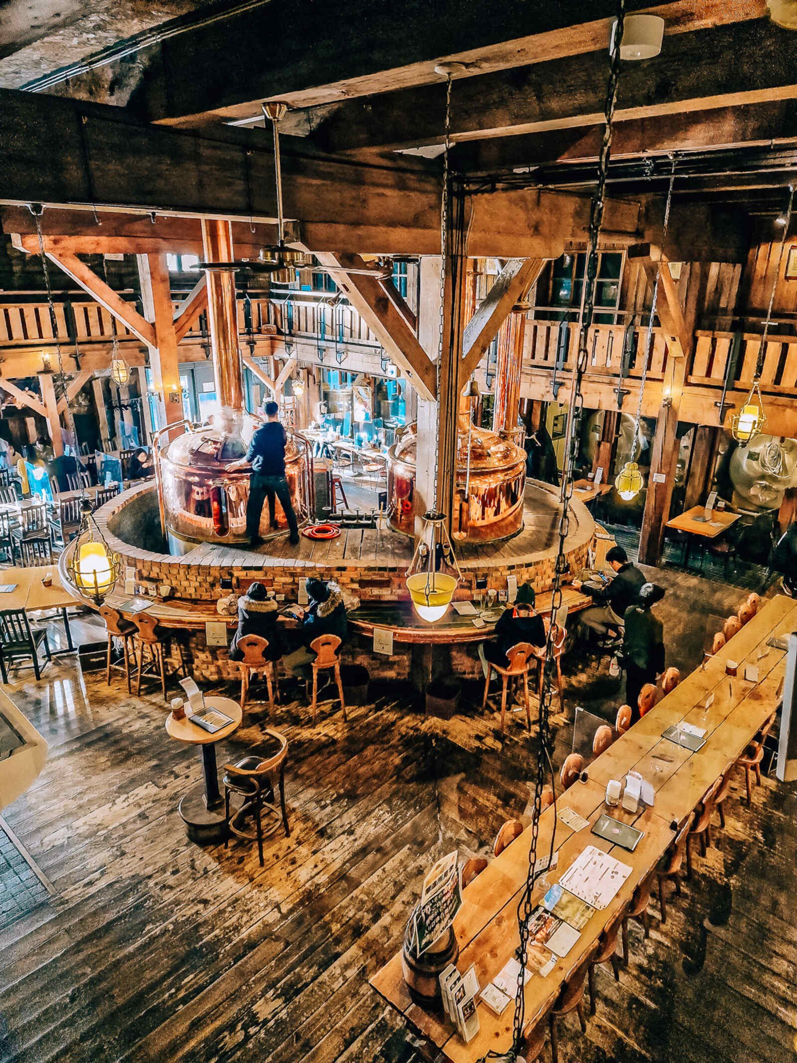 a large brewery room in a warehouse completely made of wood with seating tables and a copper beer fermentation tanks in the middle