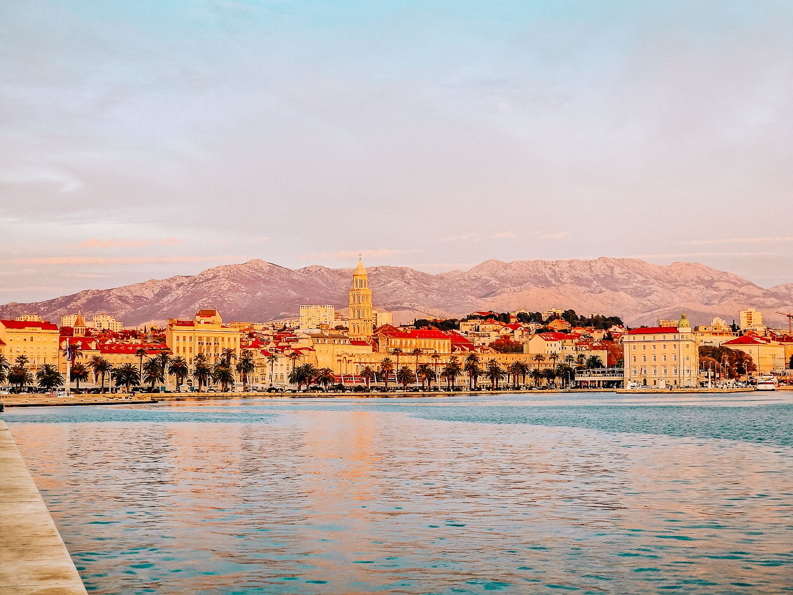 Looking across a still harbour with a town on the opposite side. Orange roof buildings and a tall stone bell tower are on the waterfront with palm trees along the water. Mountains in the background and everything has a pink glow as it's sunset