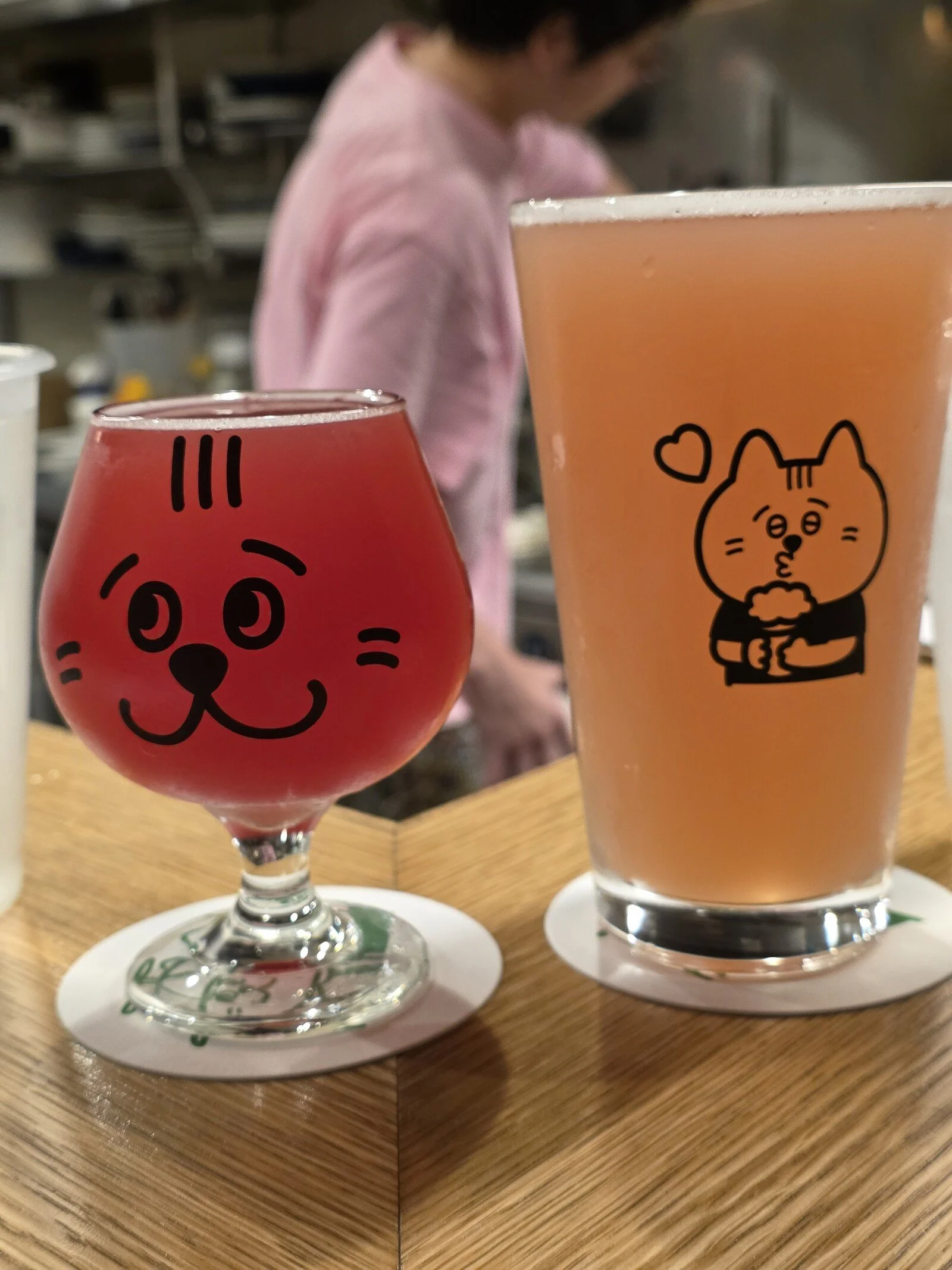 two pints of beer on a table, one is red and one is orange, both glasses have cartoon line drawings of cats on them