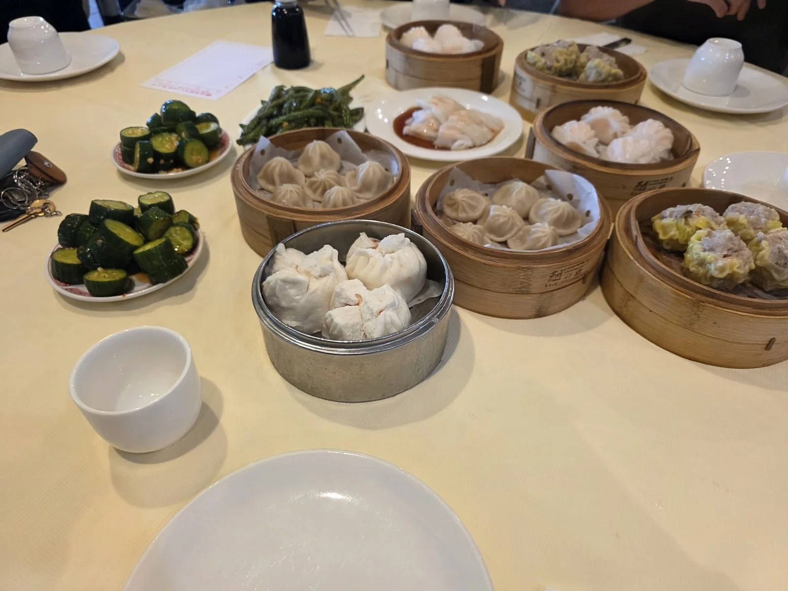 A table with 10+ wooden bowls and plates of dim sum with dumplings and buns on each and small plates of pickled cucumbers