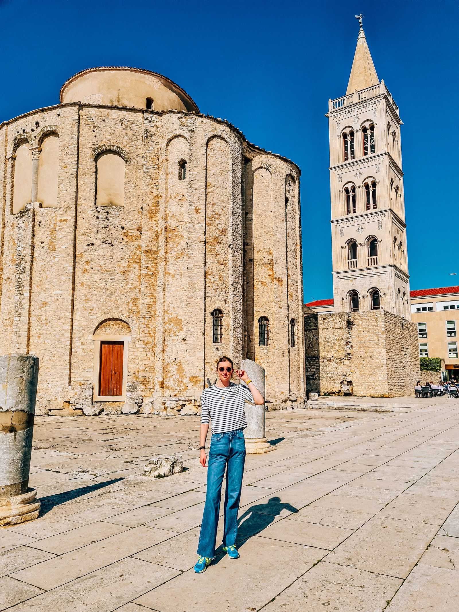 Girl in blue jeans standing infront of a large old stone circular shaped church and a stone bell tower on its right side