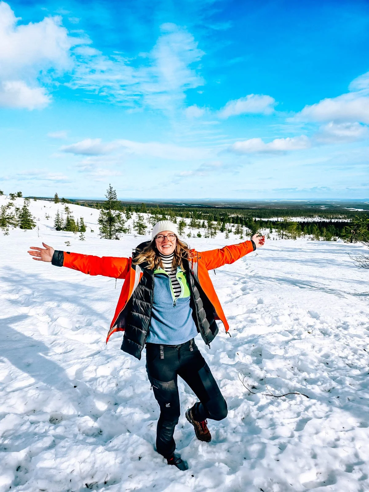 Helena in red standing in snow with her arms open. A vast snow and pine tree covered landscape is in the background